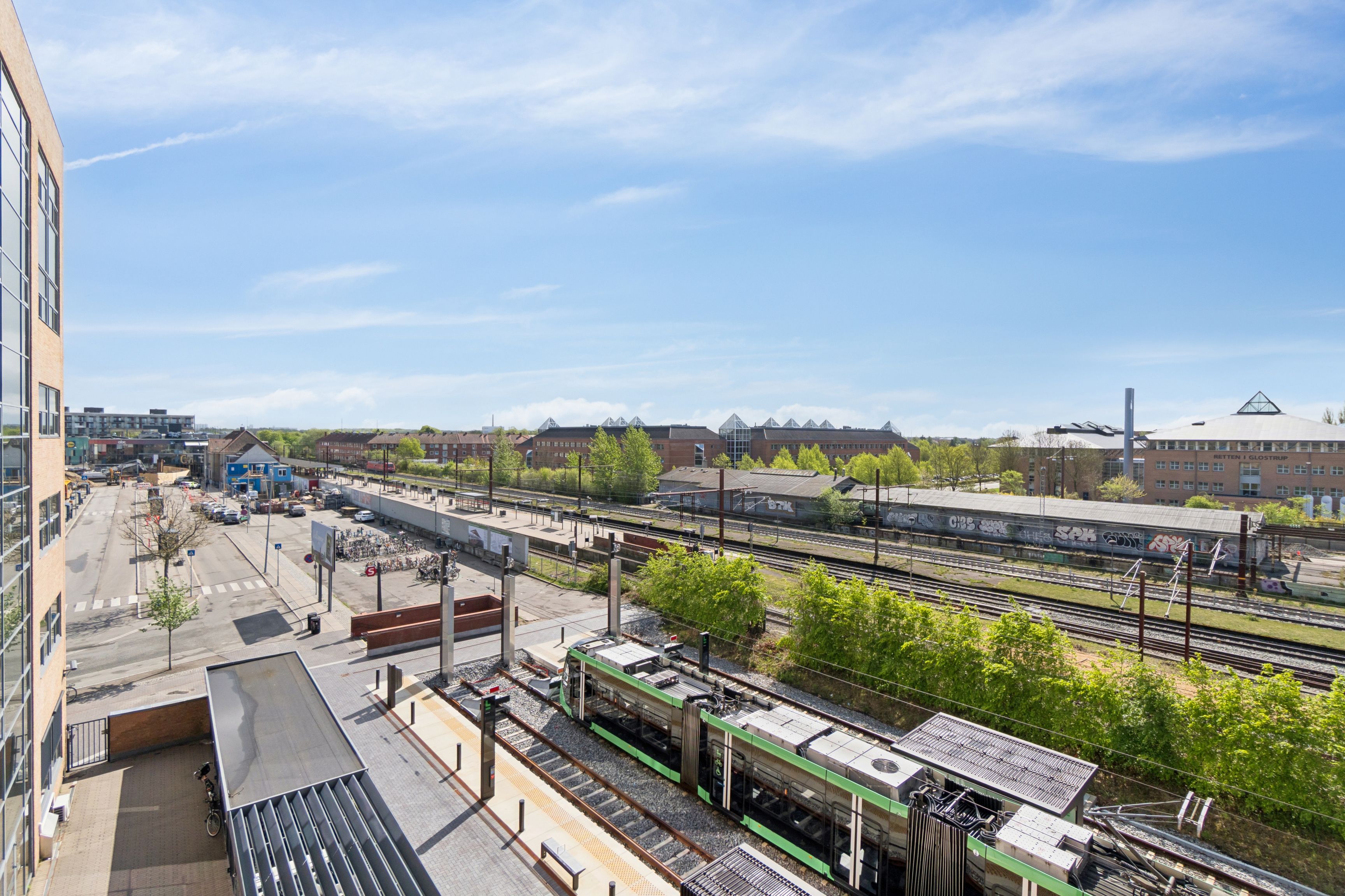 Glasvinduer med udsigt til en station med tog, togspor, busstop, parkeringsplads og grønne træer og busser i baggrunden