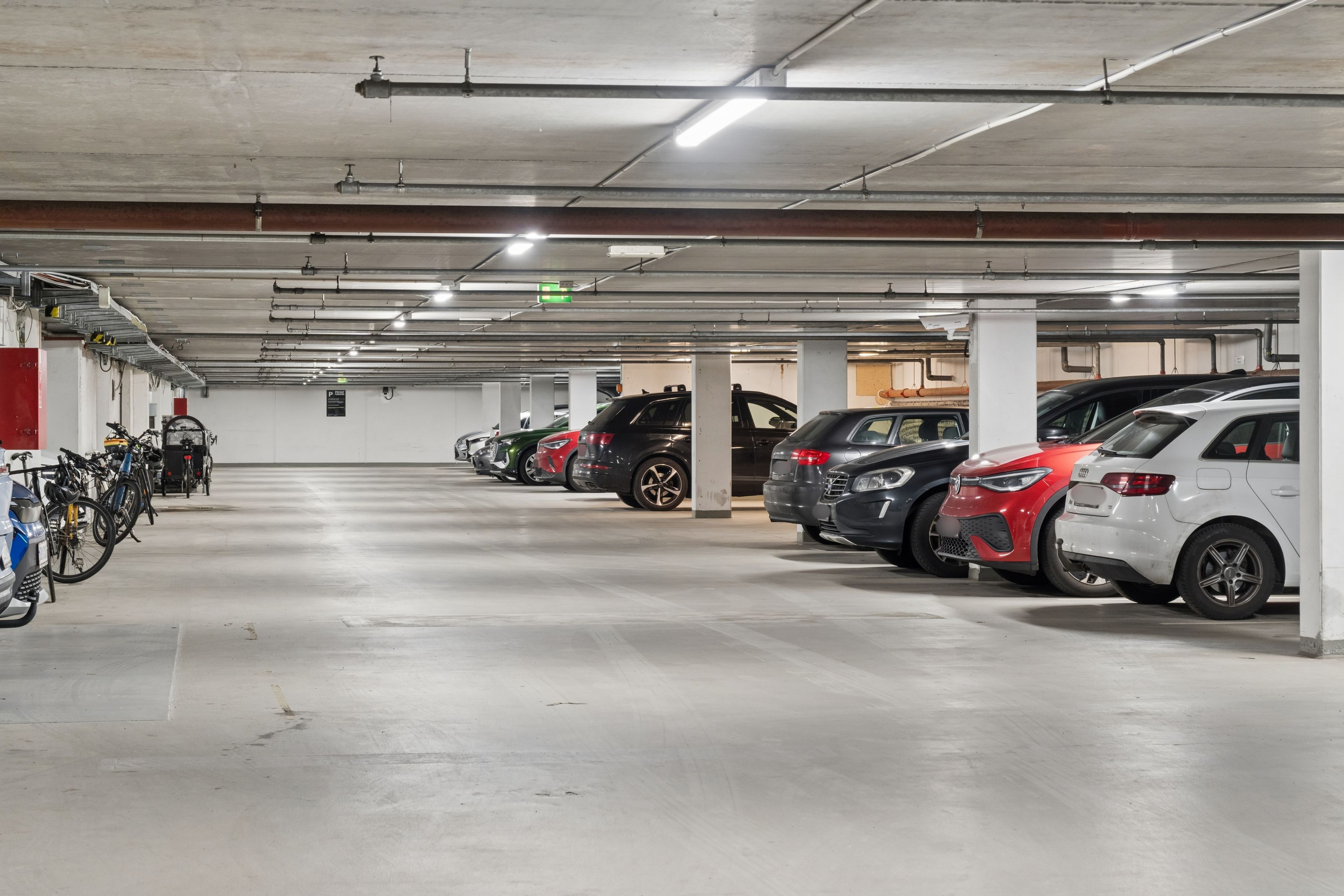 Garage med flere biler parkeret. Højt loft med lysekroner. Parkerede cykler langs væggen. Plakat med informationer og advarsler.