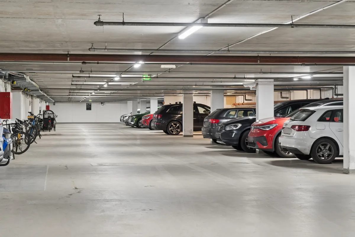 Garage med flere biler parkeret. Højt loft med lysekroner. Parkerede cykler langs væggen. Plakat med informationer og advarsler.