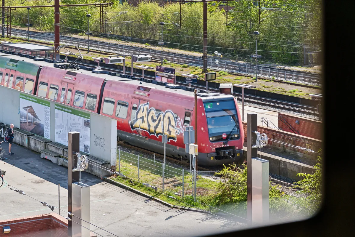 Et moderne rød tog med graffiti står ved et spor med grønne buske og træer, sammen med et gammelt hus i baggrunden