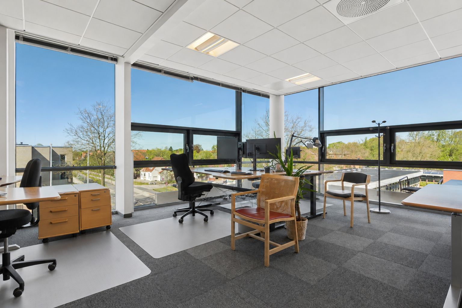 Kontor med højt lysindfald, store vinduer, moderne møbler og udsigt til grønne træer og bygninger. Lyst og rummeligt arbejdsrum med smukke udsigtsvindue.