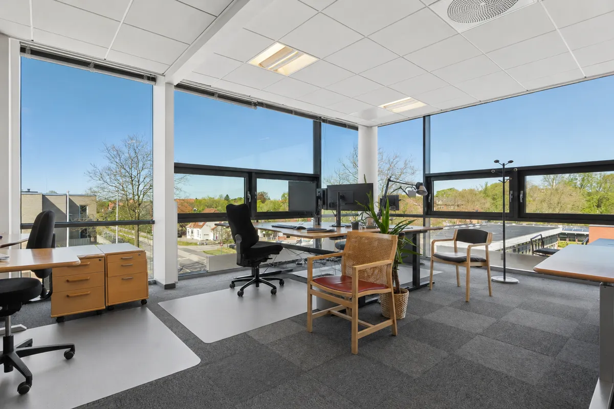 Kontor med højt lysindfald, store vinduer, moderne møbler og udsigt til grønne træer og bygninger. Lyst og rummeligt arbejdsrum med smukke udsigtsvindue.