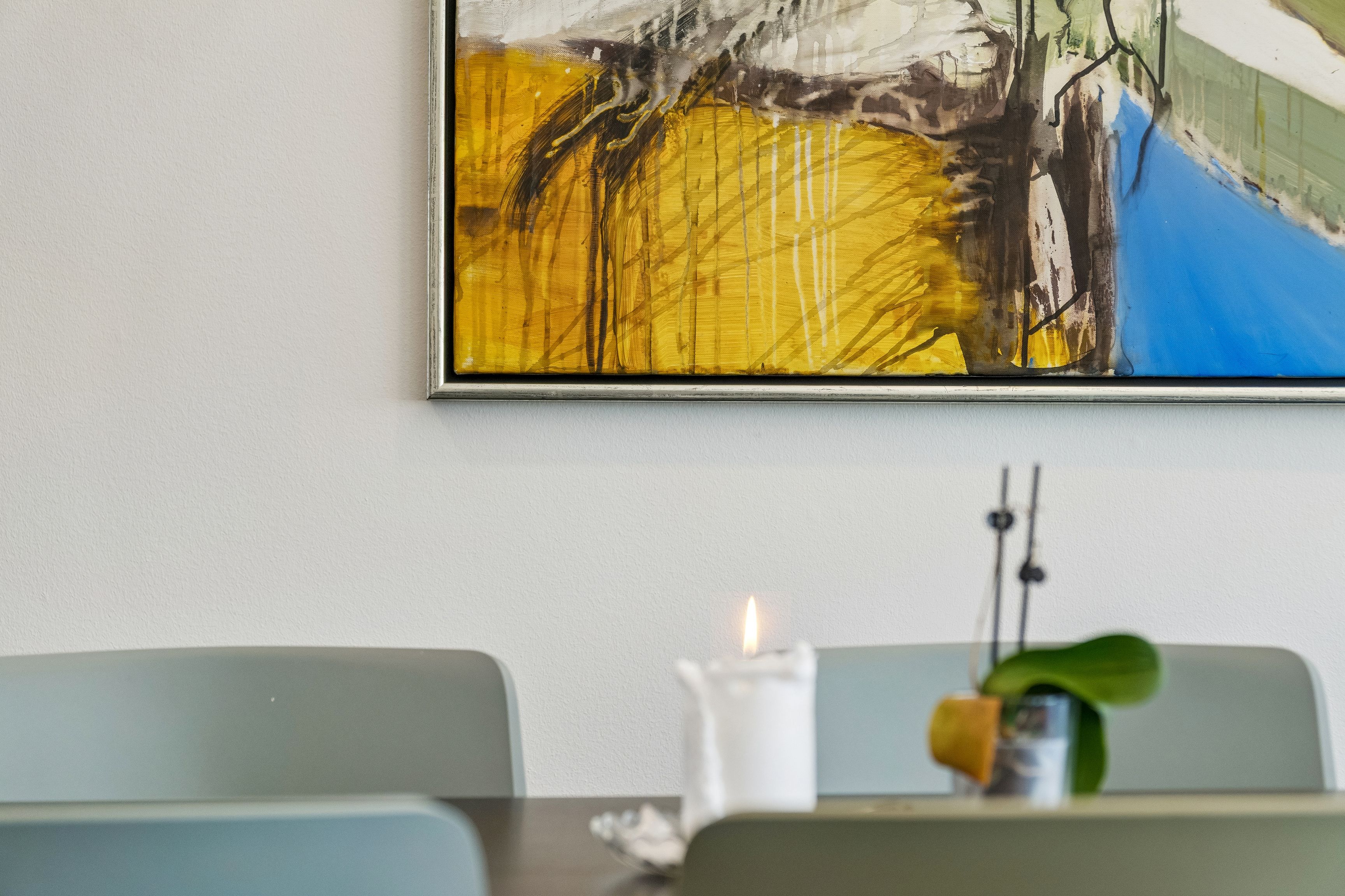 En lille stue med en stor maleri med lyse farver og en brændende stearinlys med en lille plante i en stående vase i baggrunden