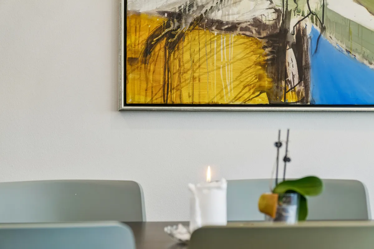 En lille stue med en stor maleri med lyse farver og en brændende stearinlys med en lille plante i en stående vase i baggrunden