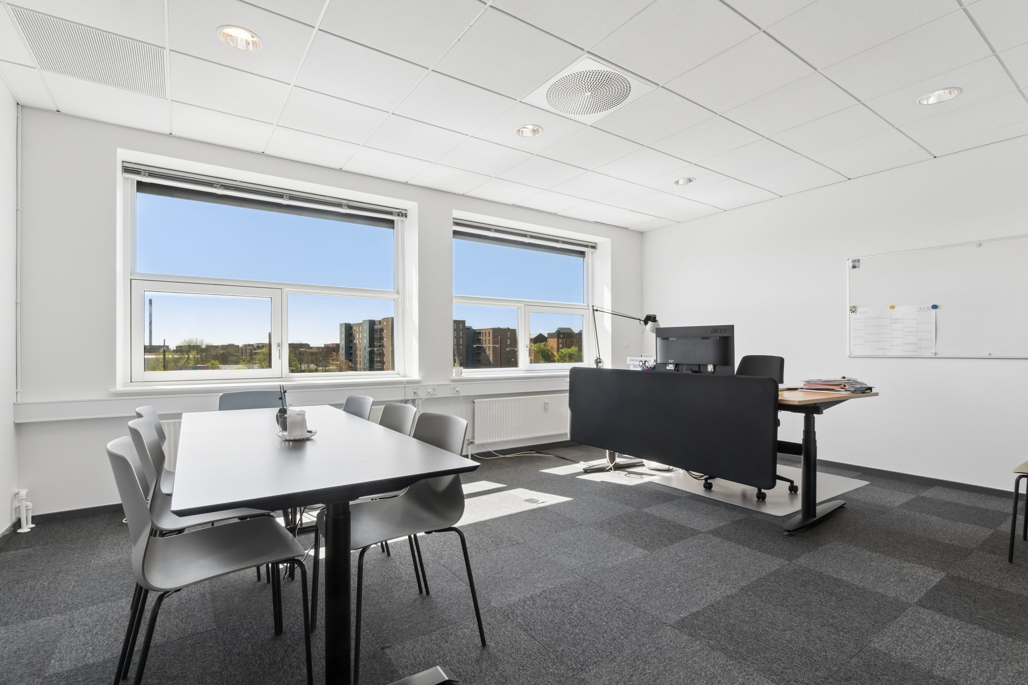 Kontor med store vinduer og lysindfald. To borde og flere stole, en hvid tavle og en sort computer. Væggene og loftet er hvide