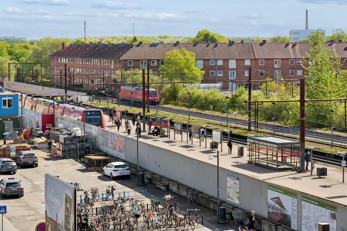 I baggrunden er et tog, der kører ind på Glostrup Station, hvor der er folk på perron og flere biler parkeret ved siden af