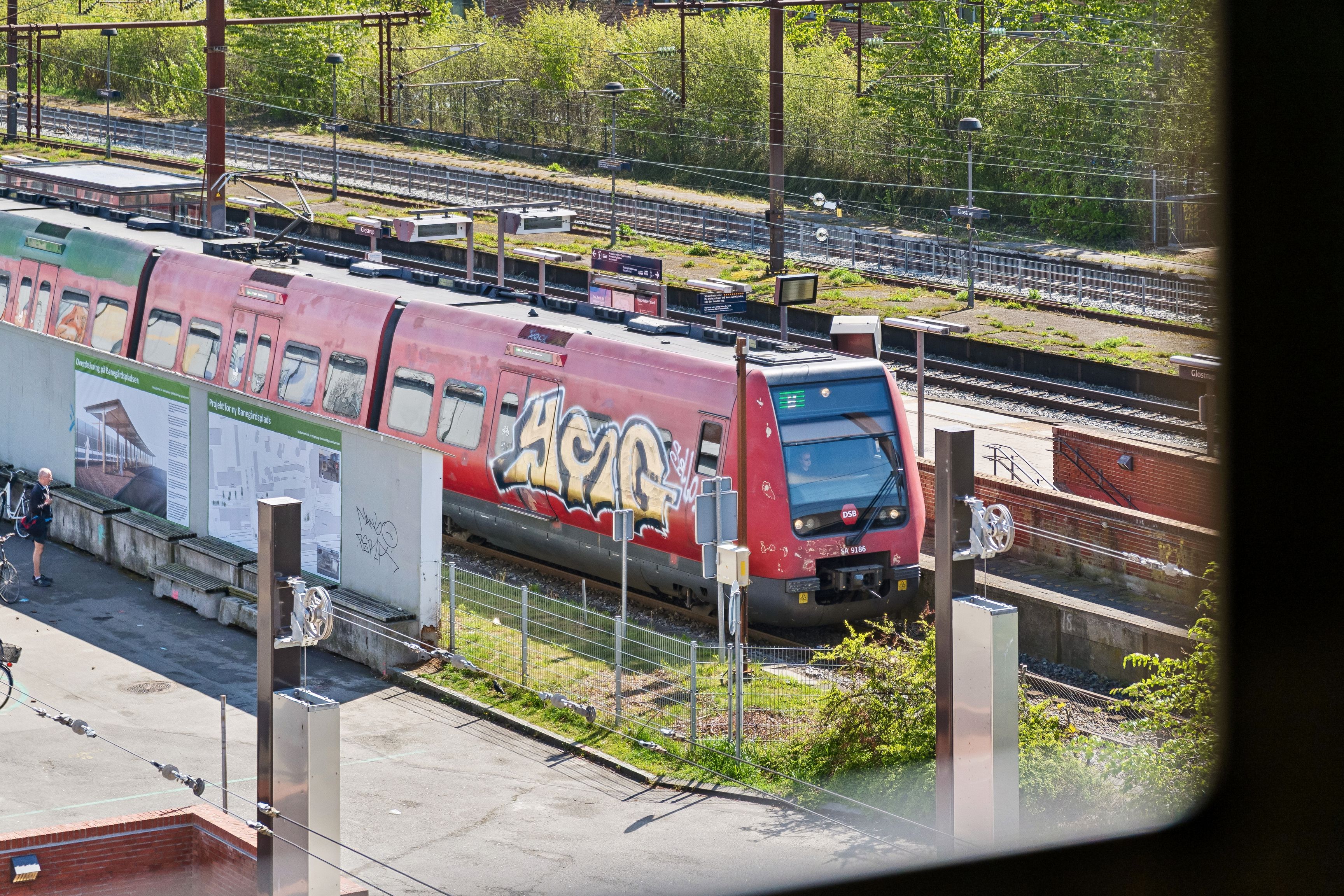 En rød togvogn står ved Glostrup Station med graffiti, mens en mand går og cykler på jernbanen, og en person sidder inde i toget