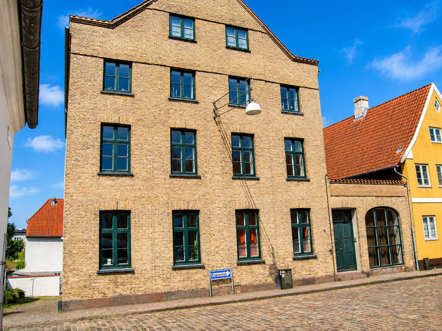Investeringsejendom på Slotsgade 21 i Haderslev. Bygningen er i gul tegl og har store vinduer og en lampestang på siden. Gulvet er brolagt og der er en stribet schild for Ensrettel.