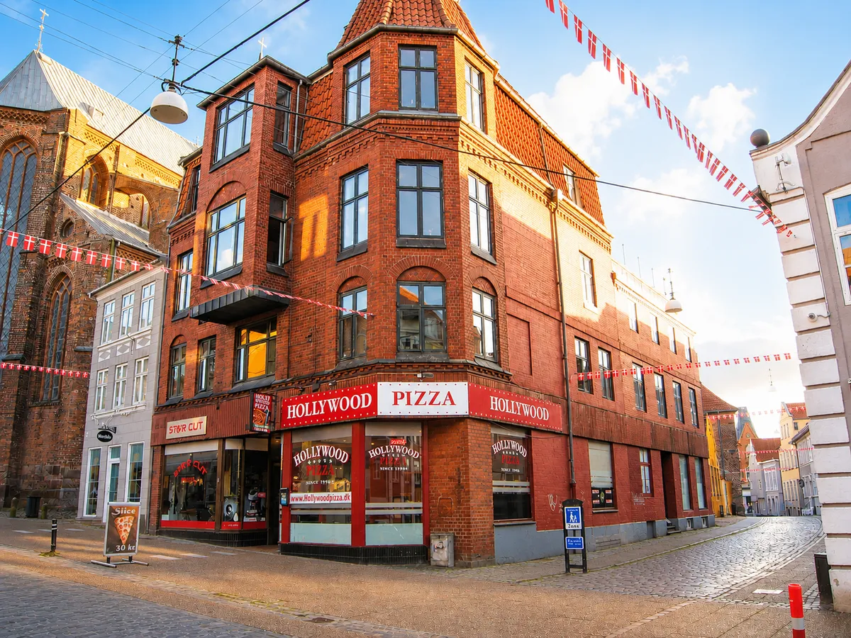 Hollywood Pizza restaurant i røde mursten. På facaden står Hollywood Pizza i rødt og hvidt. Foran er en skilt med pizza og teksten Slice 30 kr.