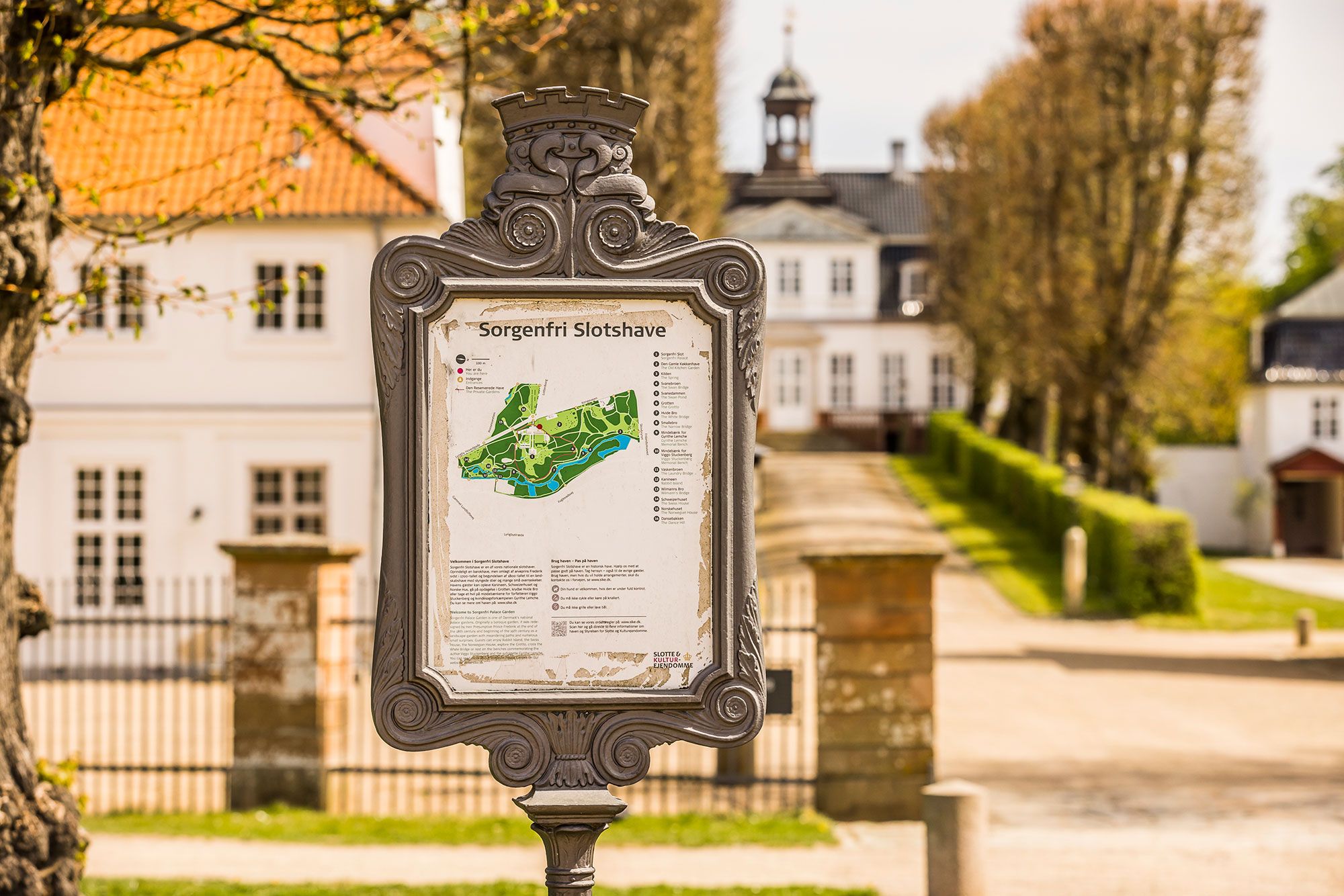 Stående søjle med kort over Sorgenfri Slothave med sø og gamle bygninger i baggrunden, som tilbyder en historisk oplevelse