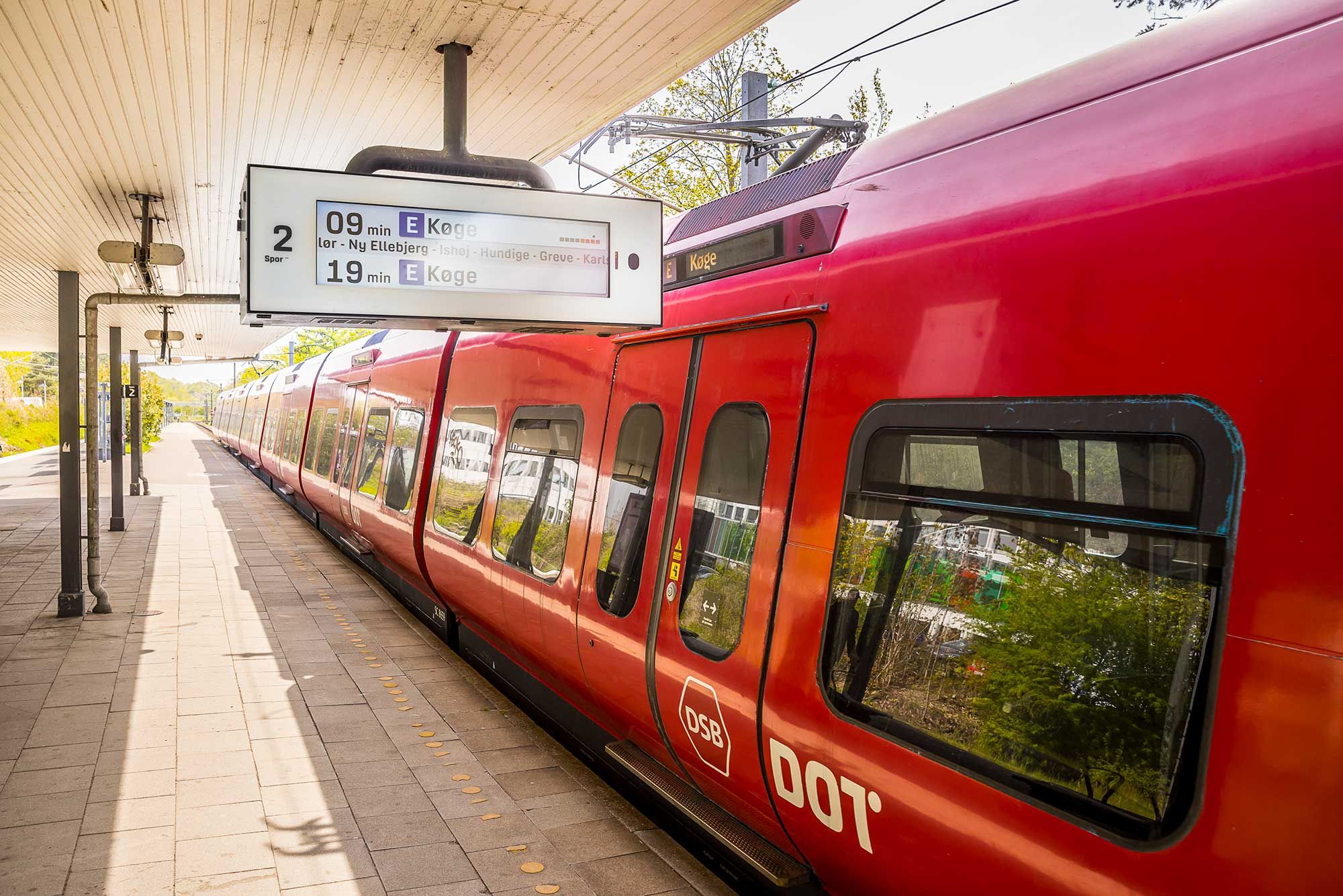 En rødt tog venter ved perronen med et skilt med tiderne til Eksjö, Nyköping og Køge. Østgående tog er ankommet og vestgående tog er ventes om 19 minutter.