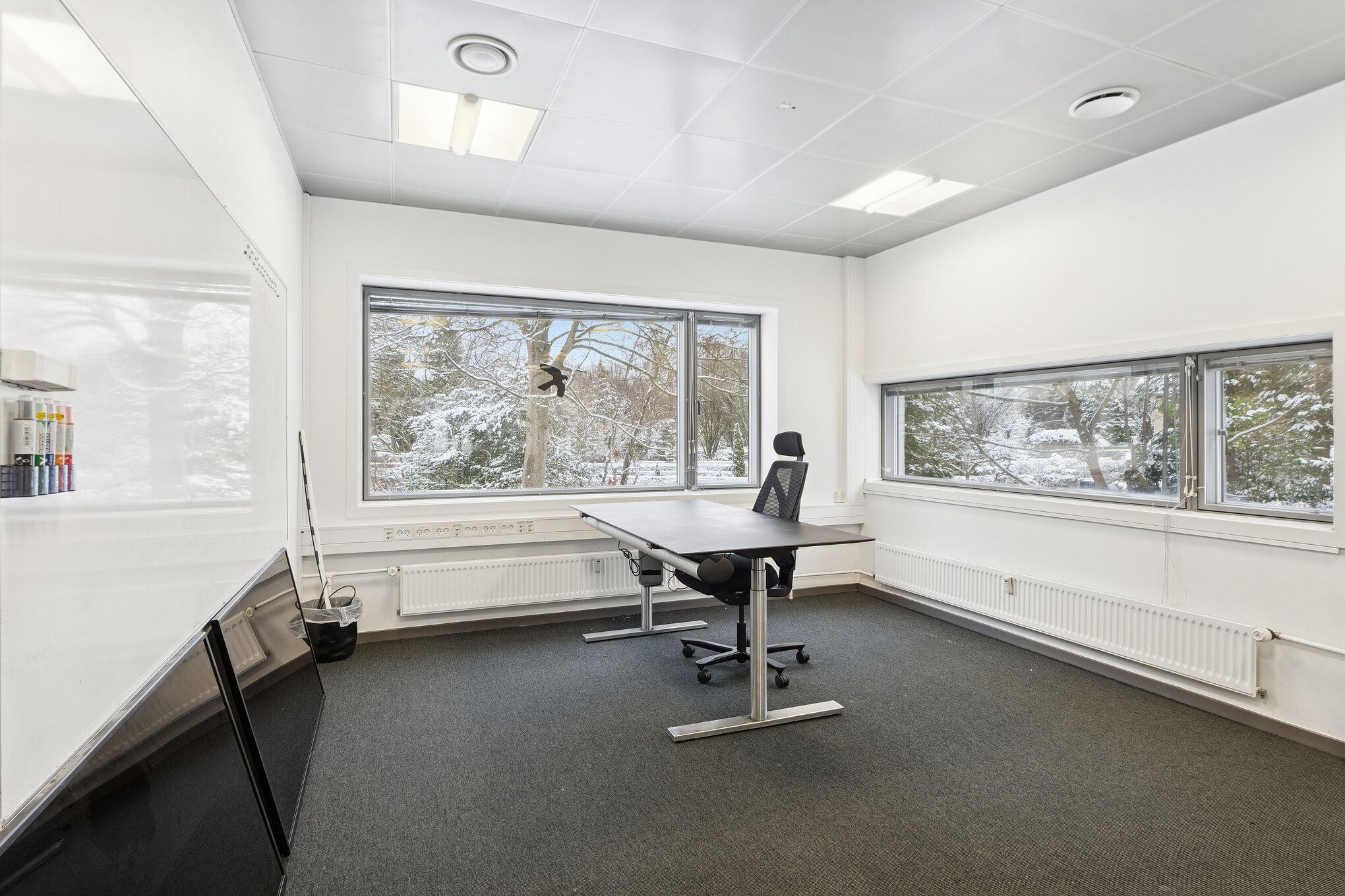 Kontor med skrivebord, stol og ruder ud mod snefremstillede træer. Vindueskiltet og skraldespand i hjørne. Pædagogiske materialer på væg og radiatorer langs væggen.