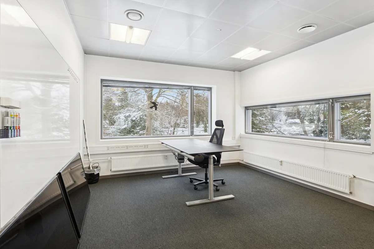 Kontor med skrivebord, stol og ruder ud mod snefremstillede træer. Vindueskiltet og skraldespand i hjørne. Pædagogiske materialer på væg og radiatorer langs væggen.