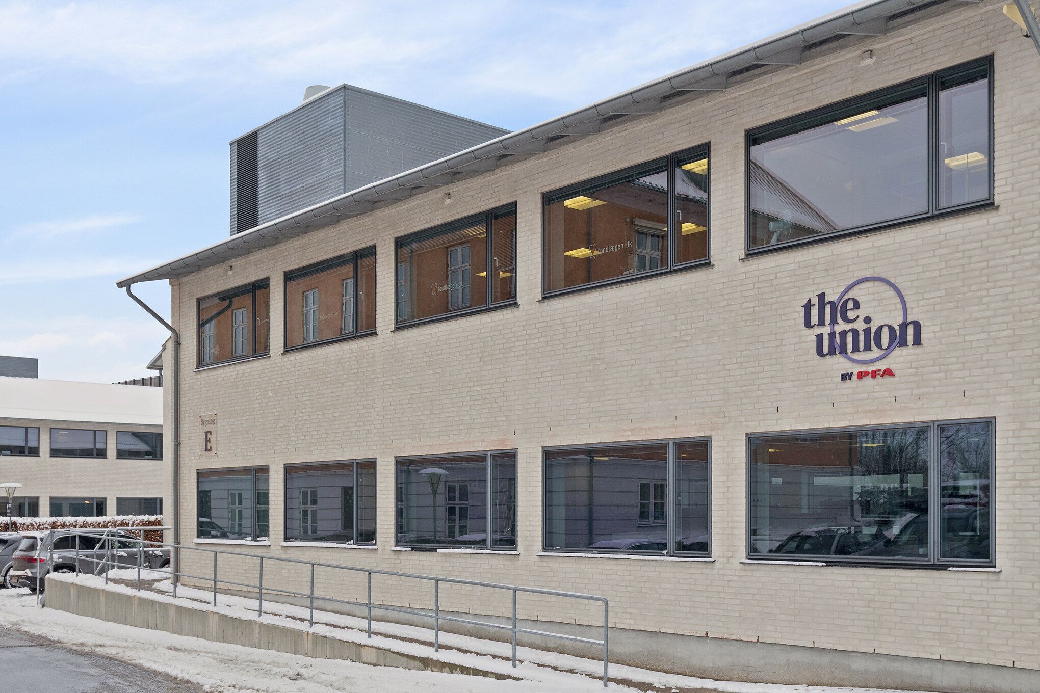 Et moderne kontorhus i Kongens Lyngby med "The Union by PFA" skrift og store vinduer. Et handicapvenligt rulkærm er installeret. Parkerede biler og sne dækker vejen foran.
