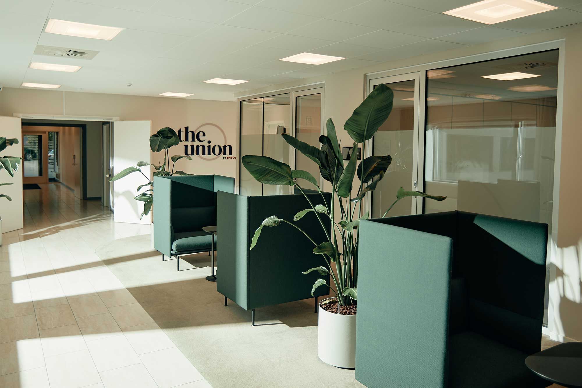 Åbent kontormiljø med grønne sofaer og store glasruder. Planter i hvide potter står i midten. Mærket "The Union" ses på væggen.