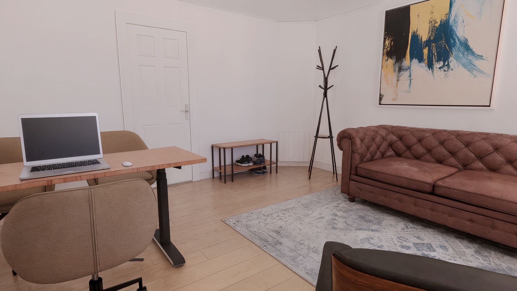Kontor med sofa og skrivebord med bærbar computer, stol og trægulv. Hjørne med træstol, ståbord og træsko. Hvid væg med maleri og hængeskab.
