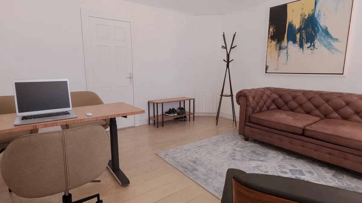 Kontor med sofa og skrivebord med bærbar computer, stol og trægulv. Hjørne med træstol, ståbord og træsko. Hvid væg med maleri og hængeskab.