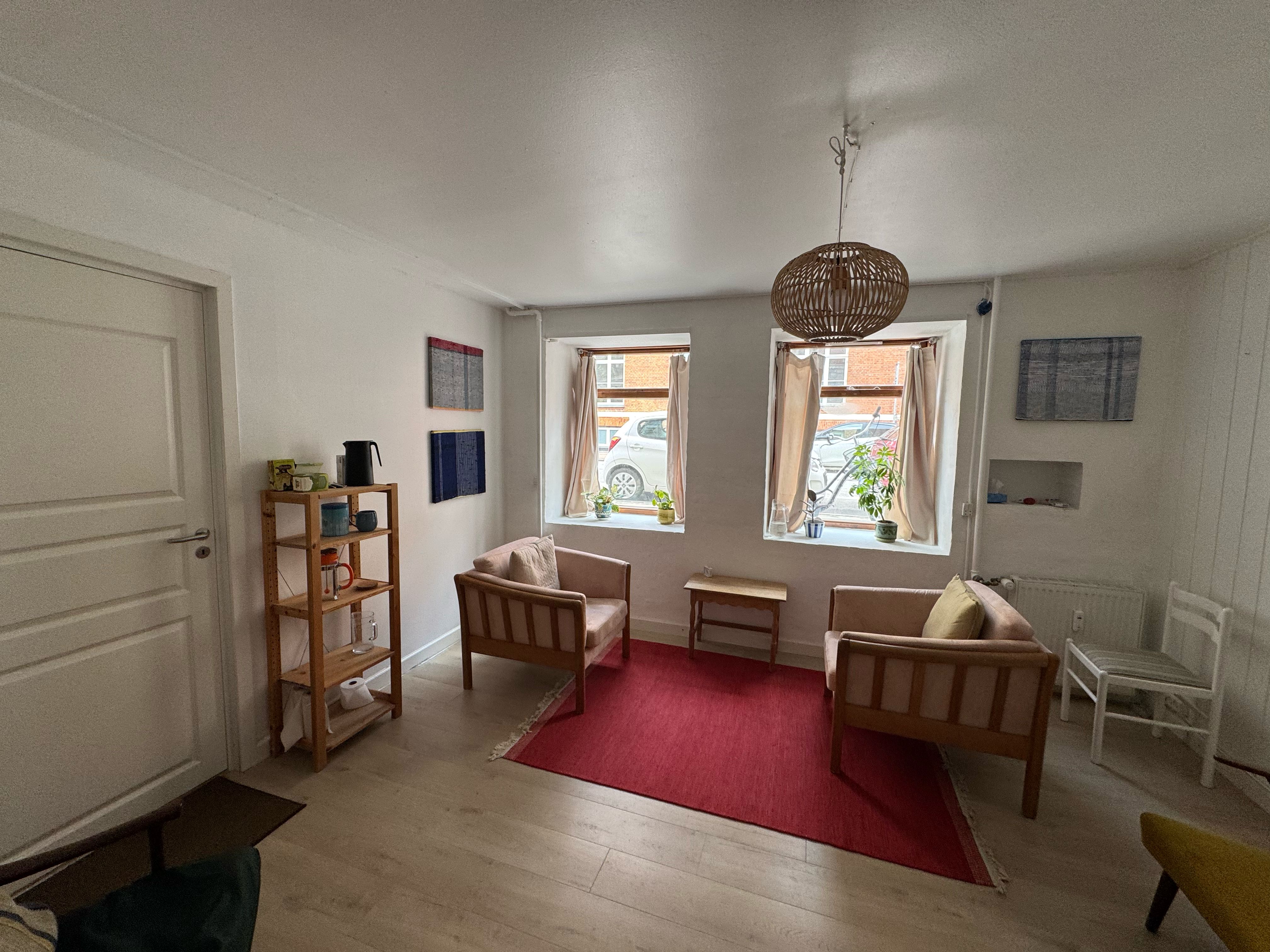 Kontor med højloft og lyse gulve, to lyse chaiselonger med puder, rødt tæppe og et bord med plante. Væggen er hvid med kunstværker og to vinduer med plante.