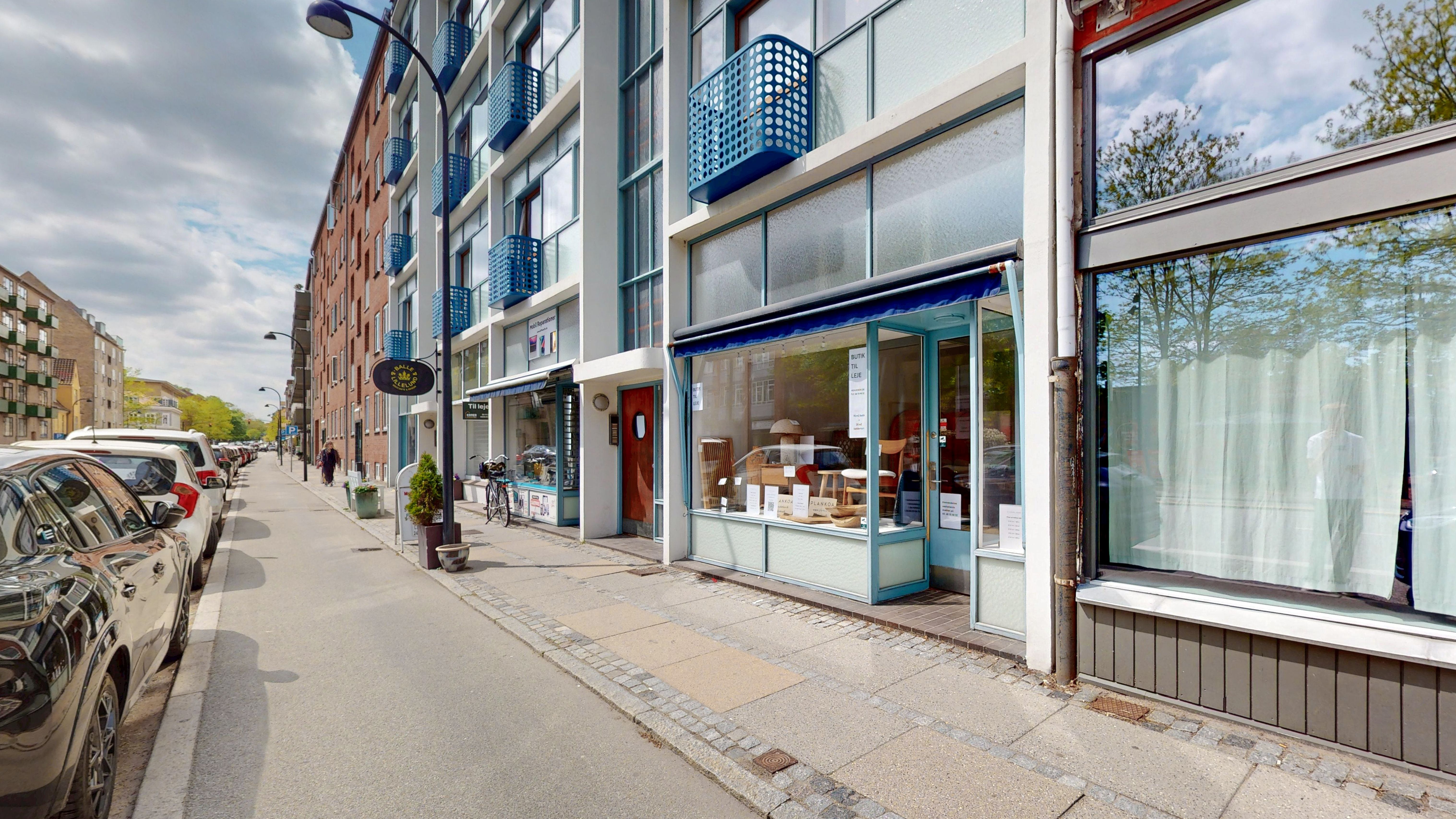 Bygade med parkerede biler, butikker, træer og lyseregn i skyerne