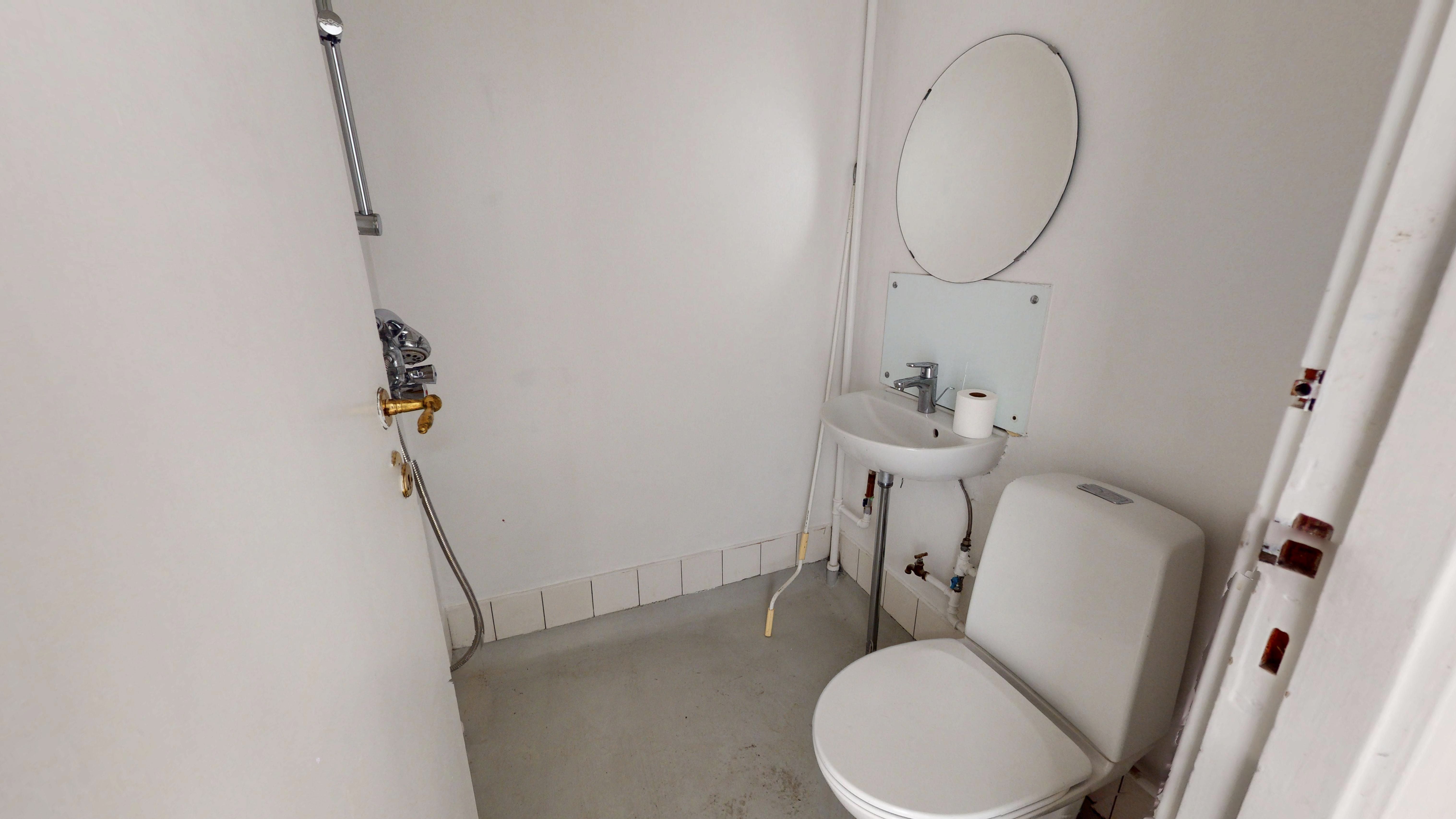 En lille, hvid badeværelse med toilet, håndvask og spejl samt et bruserhoved på væggen. Der er en lyseblå, tyk slange og to gule vandhaner.