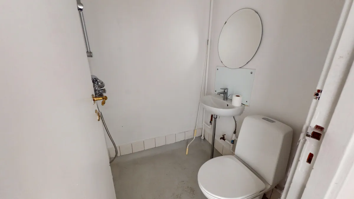 En lille, hvid badeværelse med toilet, håndvask og spejl samt et bruserhoved på væggen. Der er en lyseblå, tyk slange og to gule vandhaner.