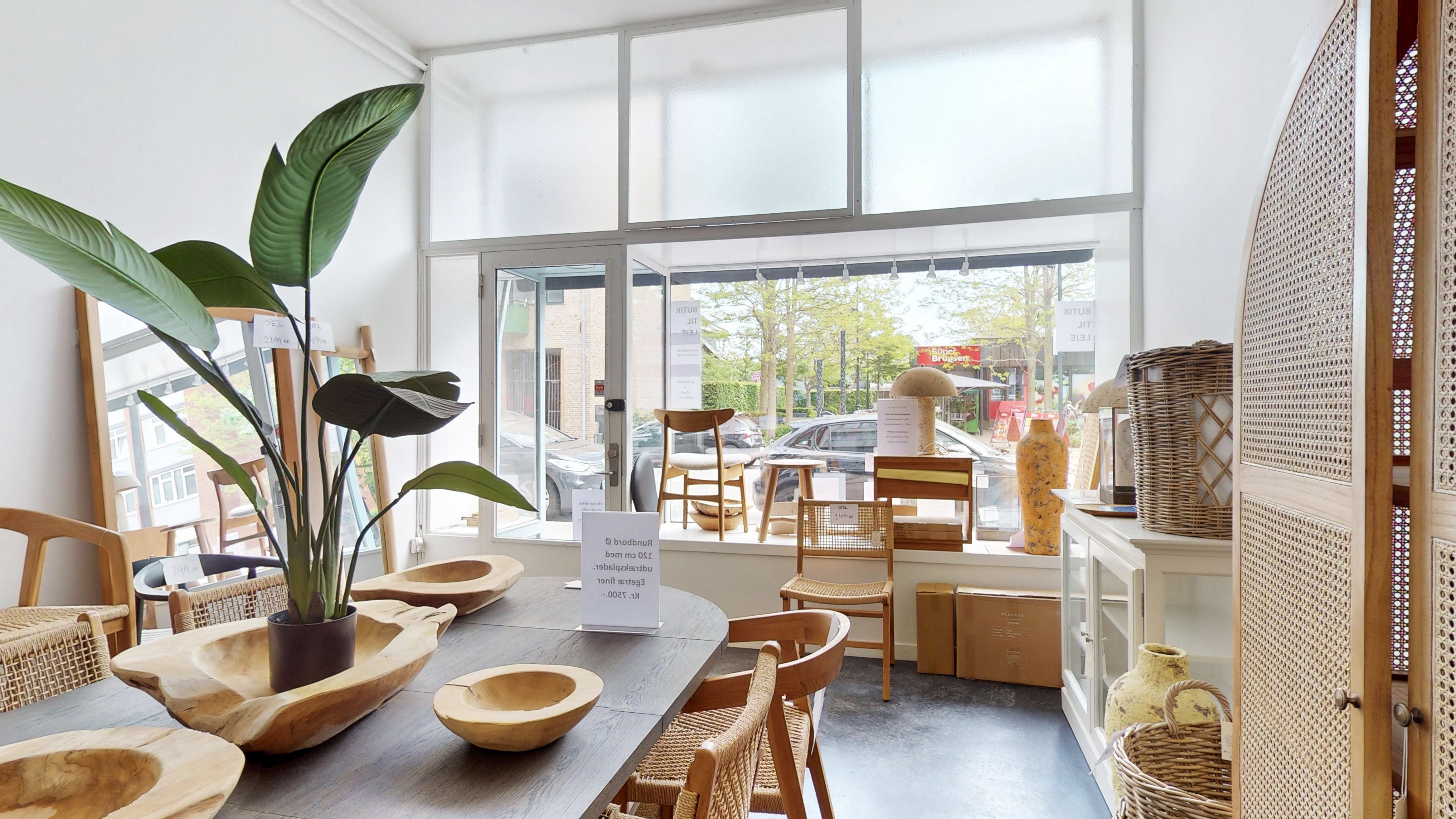 Inde i et kontorlokal er der en massiv bord, træstole og store planter. Der er også to små kurve og en vase på en høj hylde. I vinduet er der en plante og et papir med tekst.