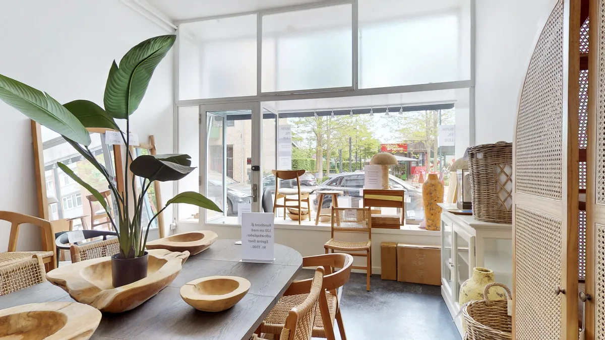 Inde i et kontorlokal er der en massiv bord, træstole og store planter. Der er også to små kurve og en vase på en høj hylde. I vinduet er der en plante og et papir med tekst.