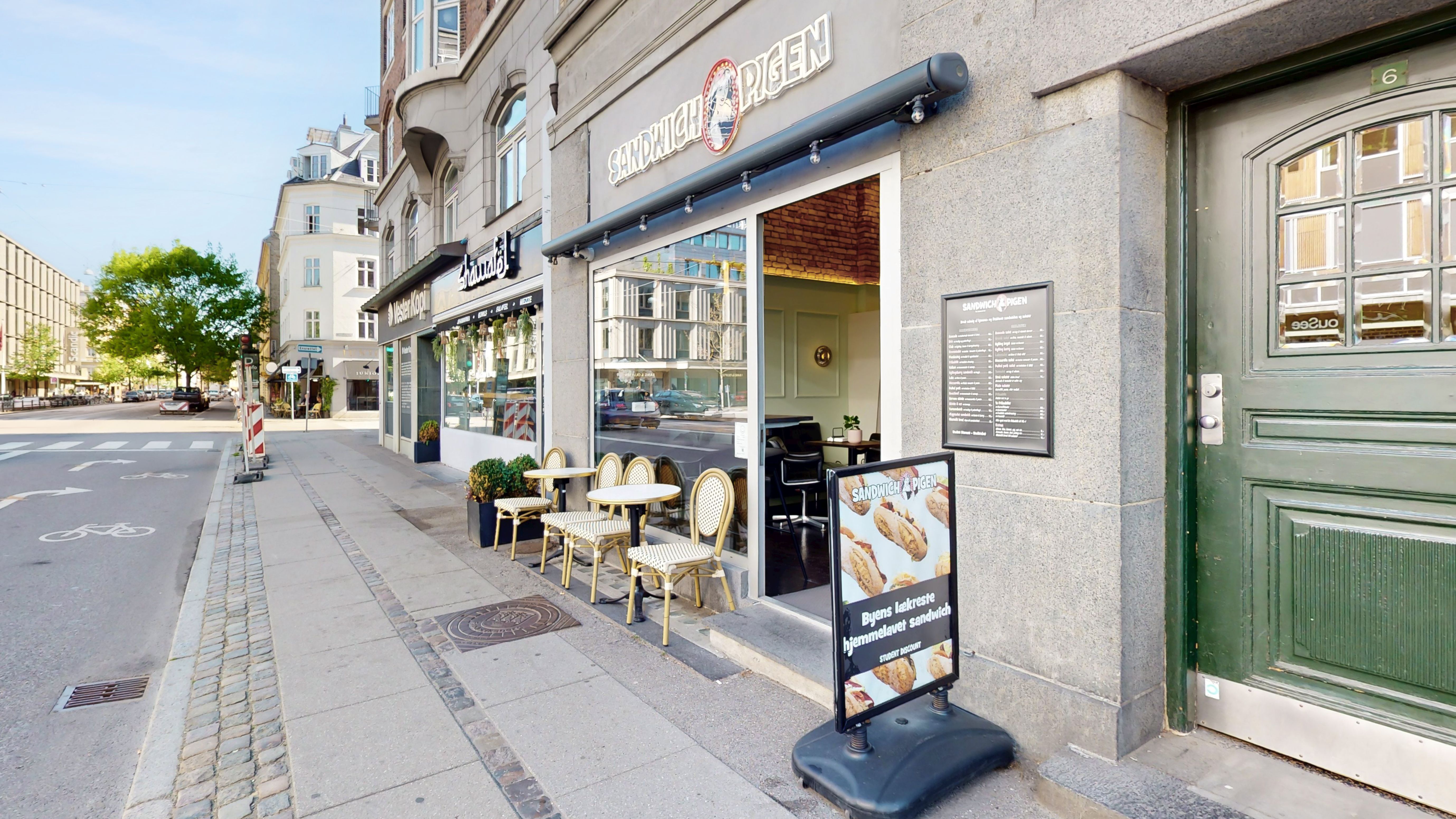 En cafe med navnet Sandwich & Pigen placeret på Falkoner Alle 6, st., 2000 Frederiksberg. Der er flere borde med stole og plantepotter udenfor. Der er tekst og billeder på skiltet foran caféen.