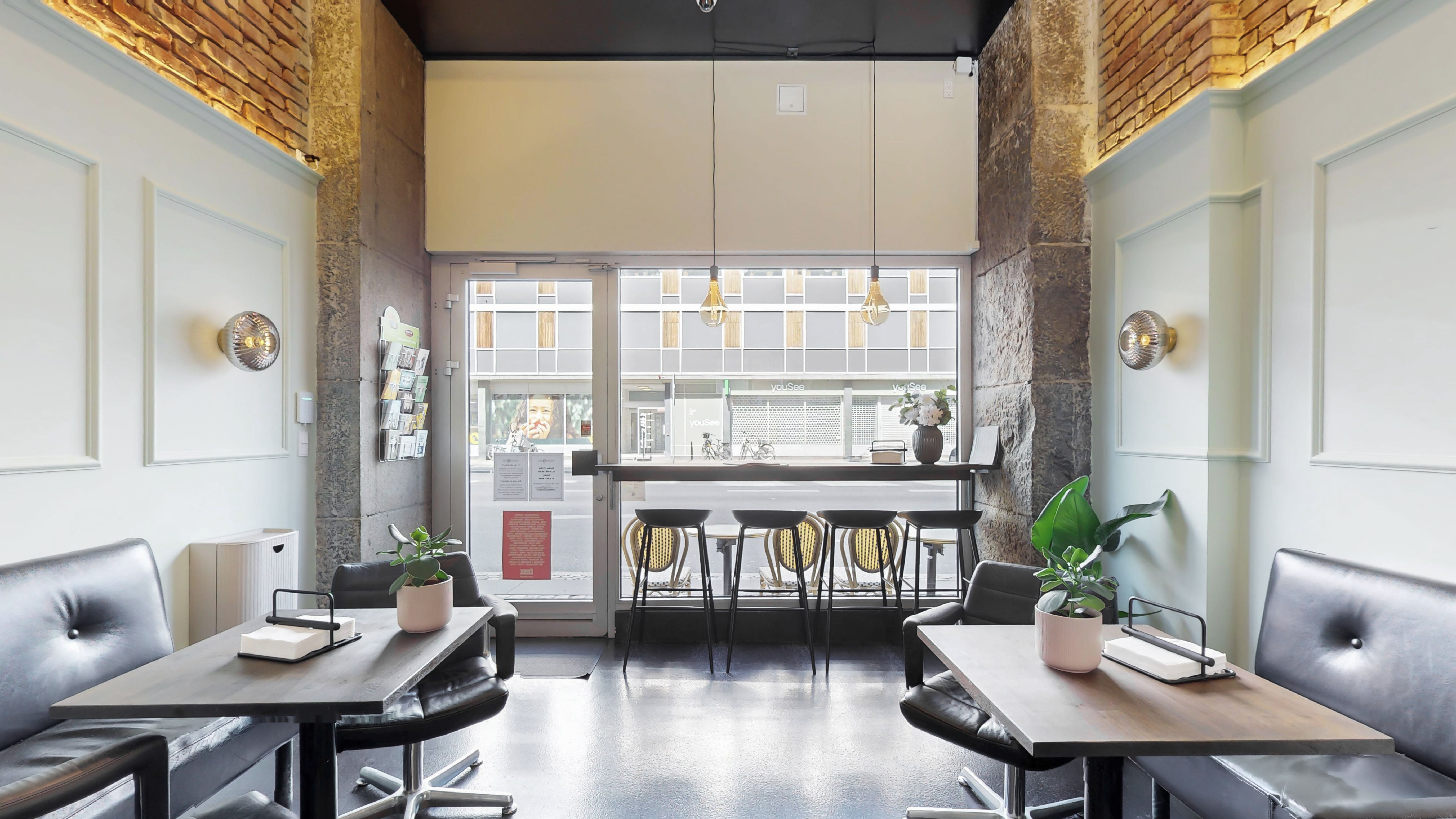 En moderne café med en stor glasdør og vinduer. Væggene er lyse og renslette, med to store sofaer og to høje stole med stolpe. Der er to høje, ovale lysekroner og en stor plante i en hvid potte på højre side
