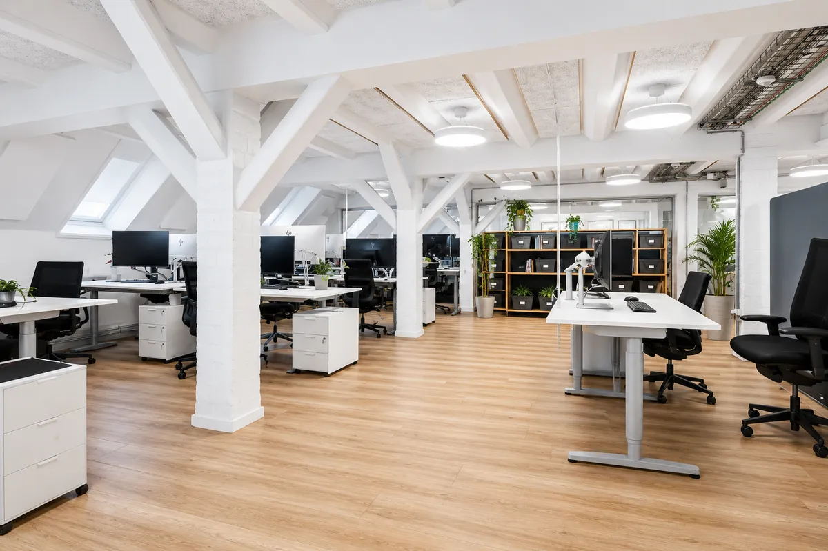 Funktionelt og lyst kontorlokal med lyse trægulve og moderne møbler. Kontoret er indrettet med skriveborde, stole og plantegrønt. Loftet er højt med bjælkeloft og loftsvinduer.