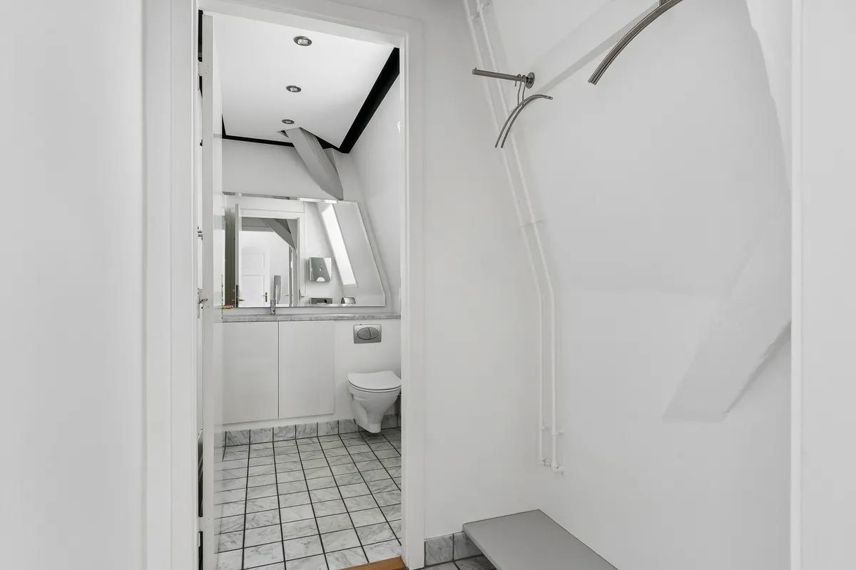 En hvid, minimalistisk badeværelse med toilet, spejl og vask, hvor hjørner og vægge er dekoreret med marmorfliser og en enkelt tørretumbler