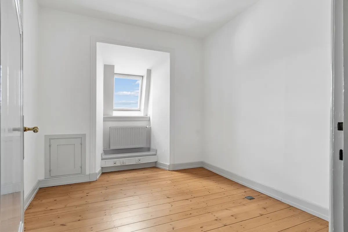 Kontor med højloft, lyse skyer, hvid væg, trægulv, radiator, vindue og dør med guldfarvet håndtag