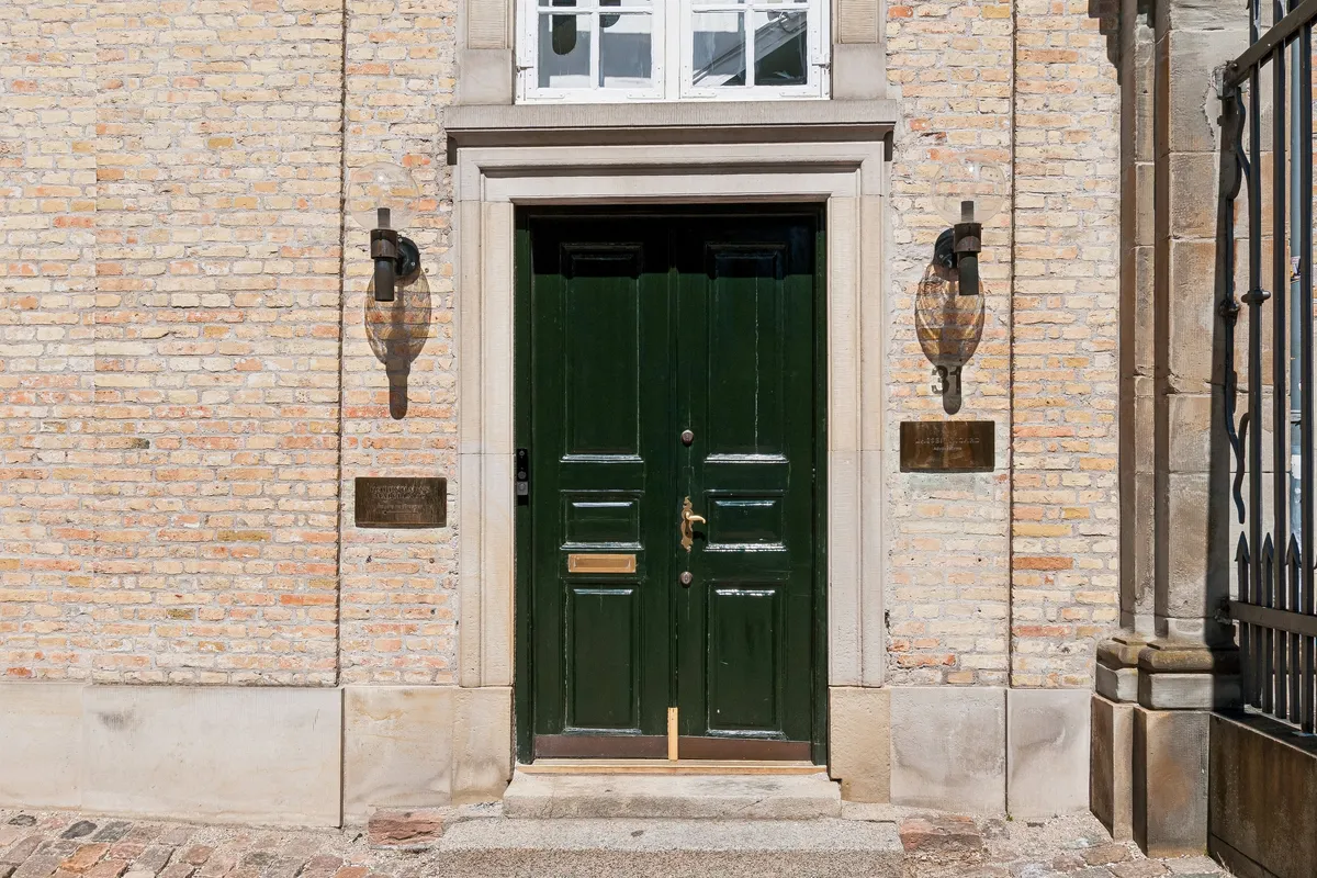 En grøn dør med lysekroner på hver side og et postnummerfelt samt en skråt liggende plaket