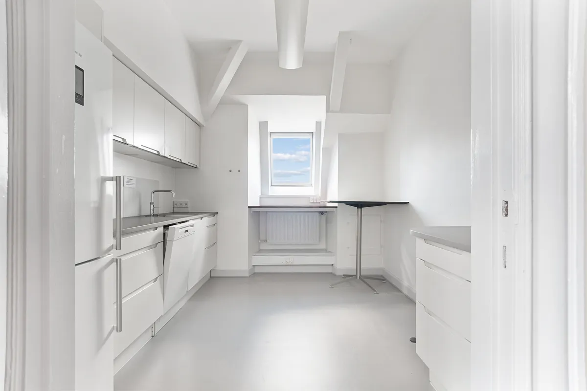 Minimalistisk hvid køkken med hvide skabe, højloft, hvid loftsbjælke, vindue med udsigt, hvid radiator, stol og hvidt køleskab