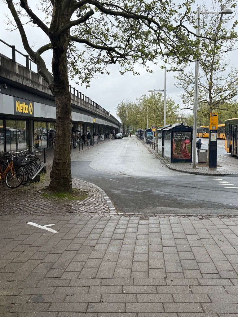 Lejlighed med udsigt til Jernbanepladsen i Kongens Lyngby, hvor busser og cykler er parkeret, og der er et skovklædt område med busstoppested og reklameskilte