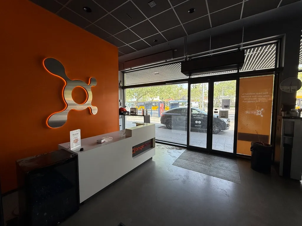 Fitnesslokale til leje ved Lyngby Station med stor vinduesfront, lyse orange vægge, moderne reception med infoskilt og indgang til loftsområde