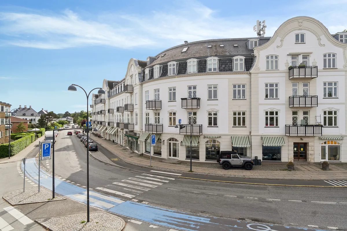 Lejlighedsbygning med balkoner og vinduer mod gaden, to biler parkeret udenfor, vej med cykelsti og parkeringspladser, fortov med grønne træer og buske, solrig himmel med skyer
