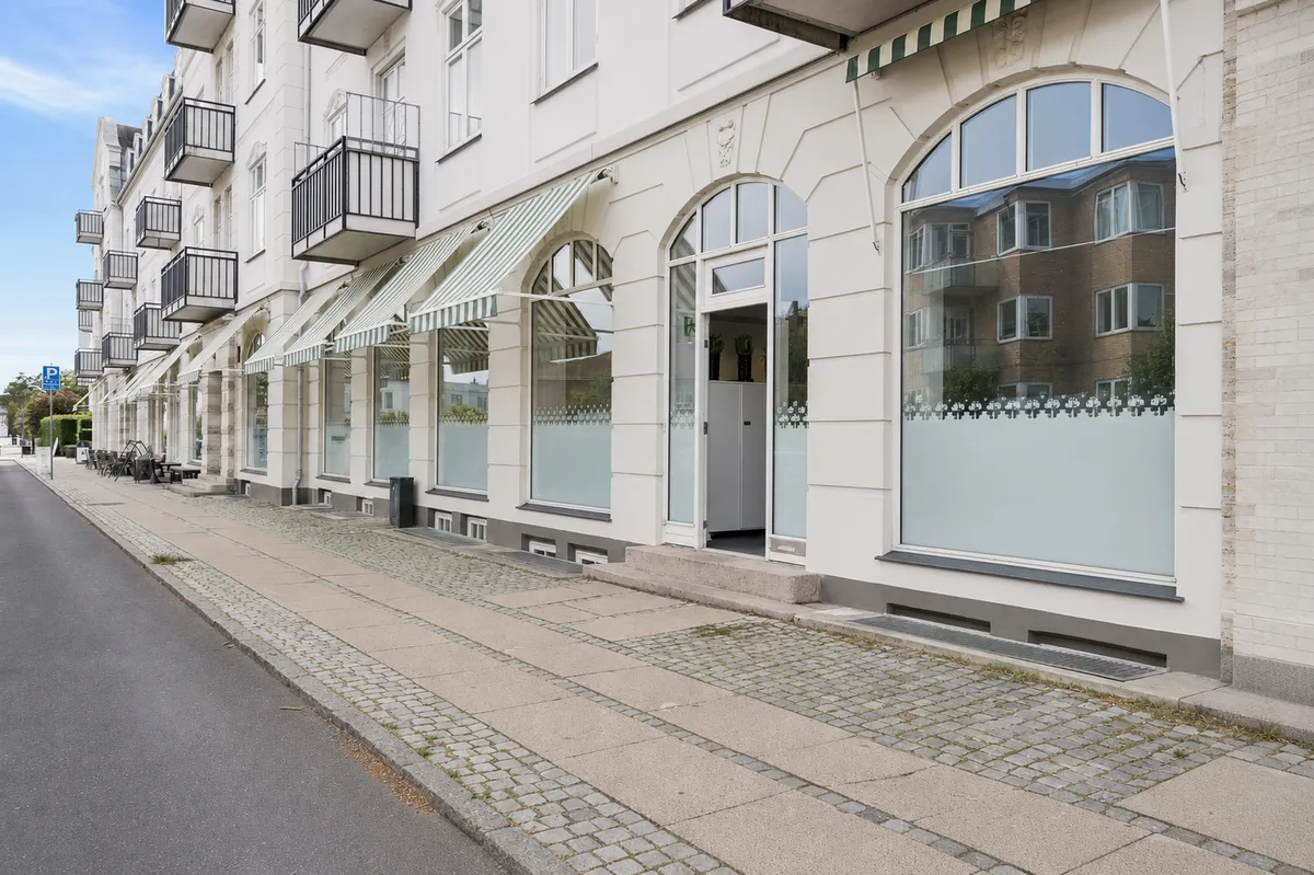 Lyst byggede bolig med store glasruder og grønne vægvindueskærme. Entré med skraldkurv og åben dør. Vej med brolægning og parkeringsplads.