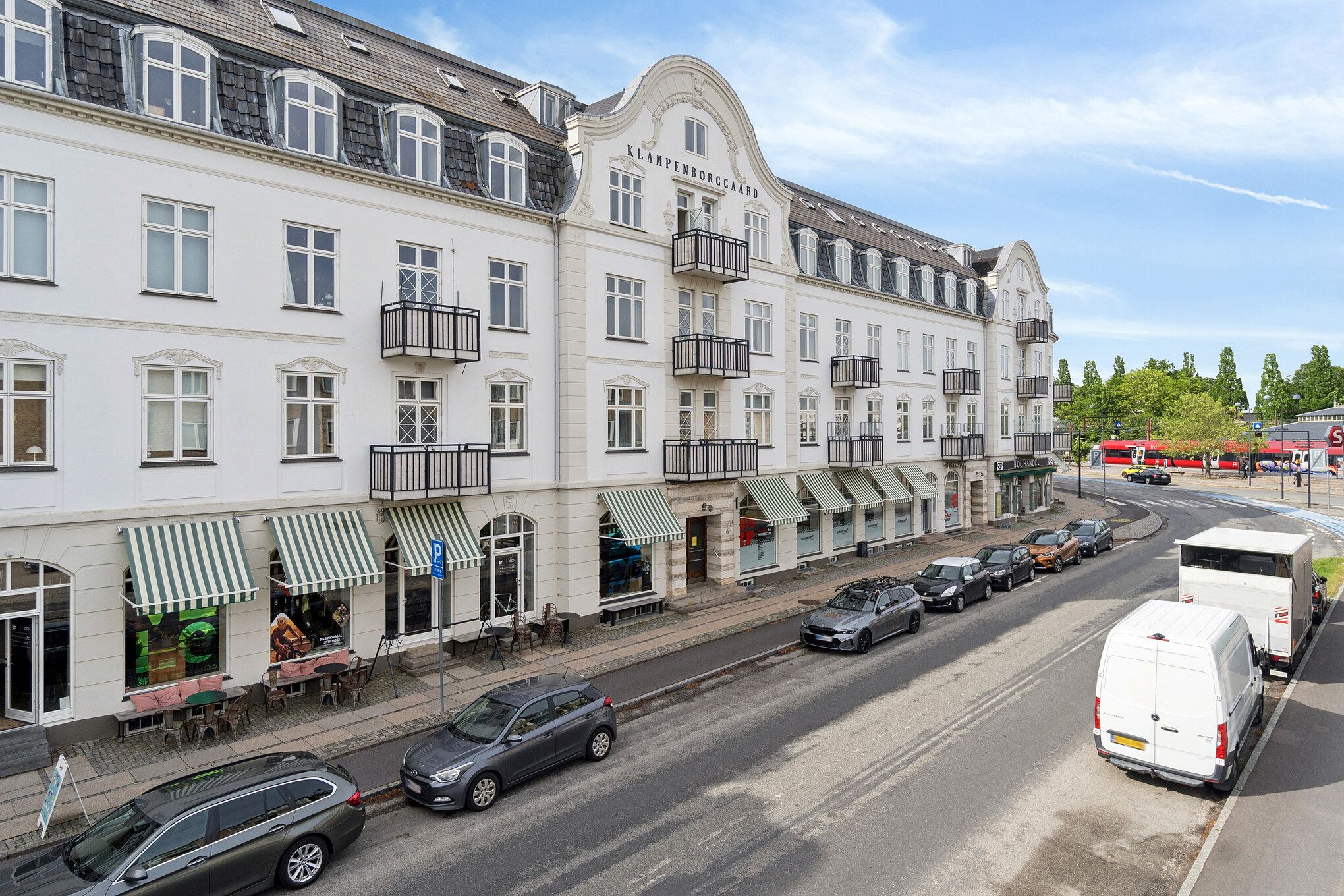 Store butikker med caféer og terrasser på Dyrehavevej 4-6 i Klampenborg. Køretøjer er parkeret langs gaden, og en sporvogn kører i baggrunden.
