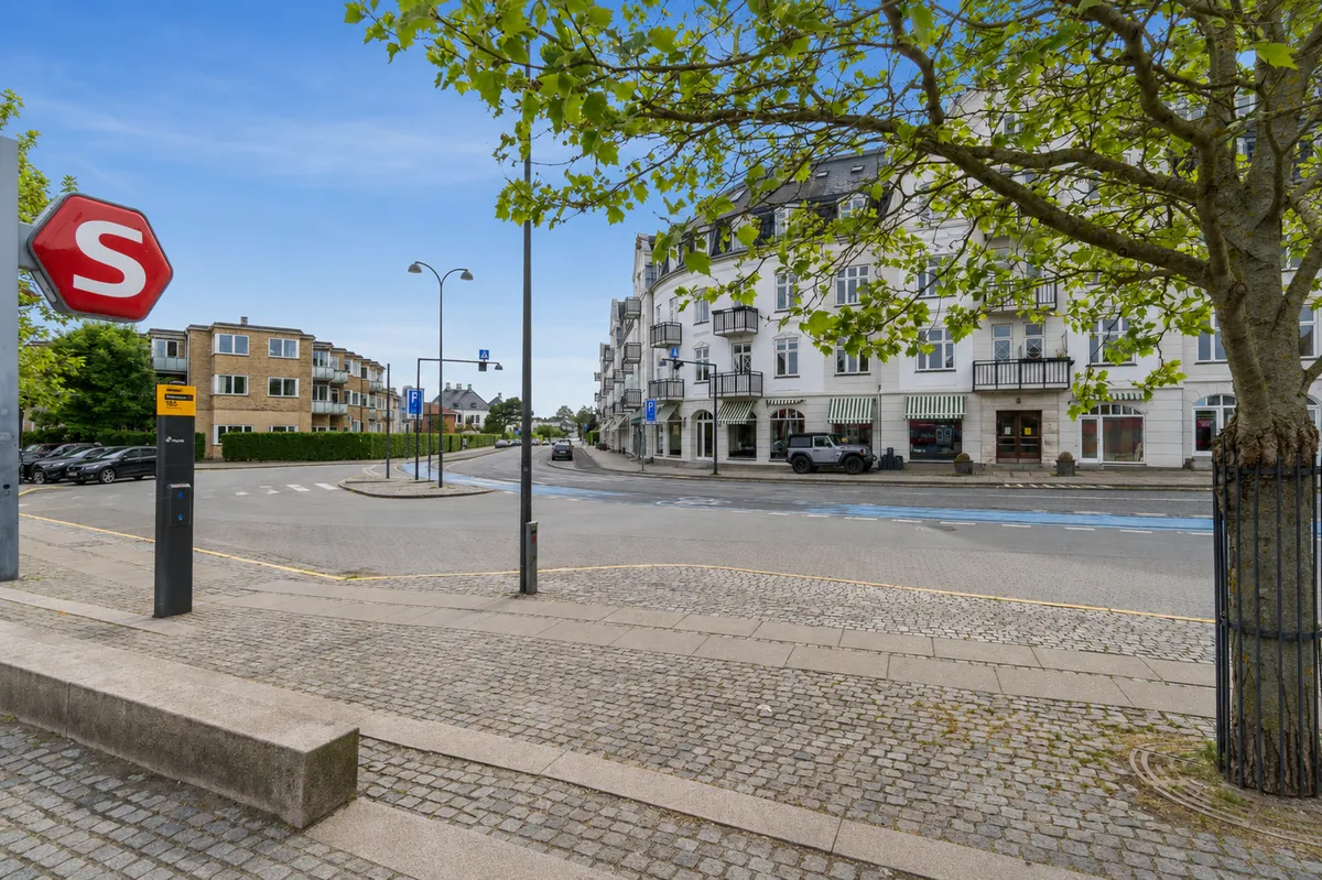 Sted med terrasse og stopskilt, hvor biler er parkeret, sammen med træer og en bygning med terrasser og parkeringsplads