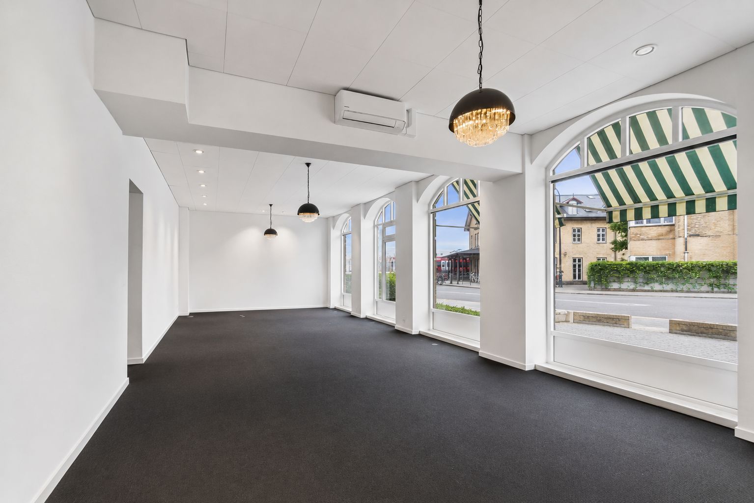 Lyst og indbydende lokale med store bueformede vinduer, højt til loftet, moderne loftsbelysning og stilren indretning. En stor lysekron og en klimaanlæg øverst i loftet.