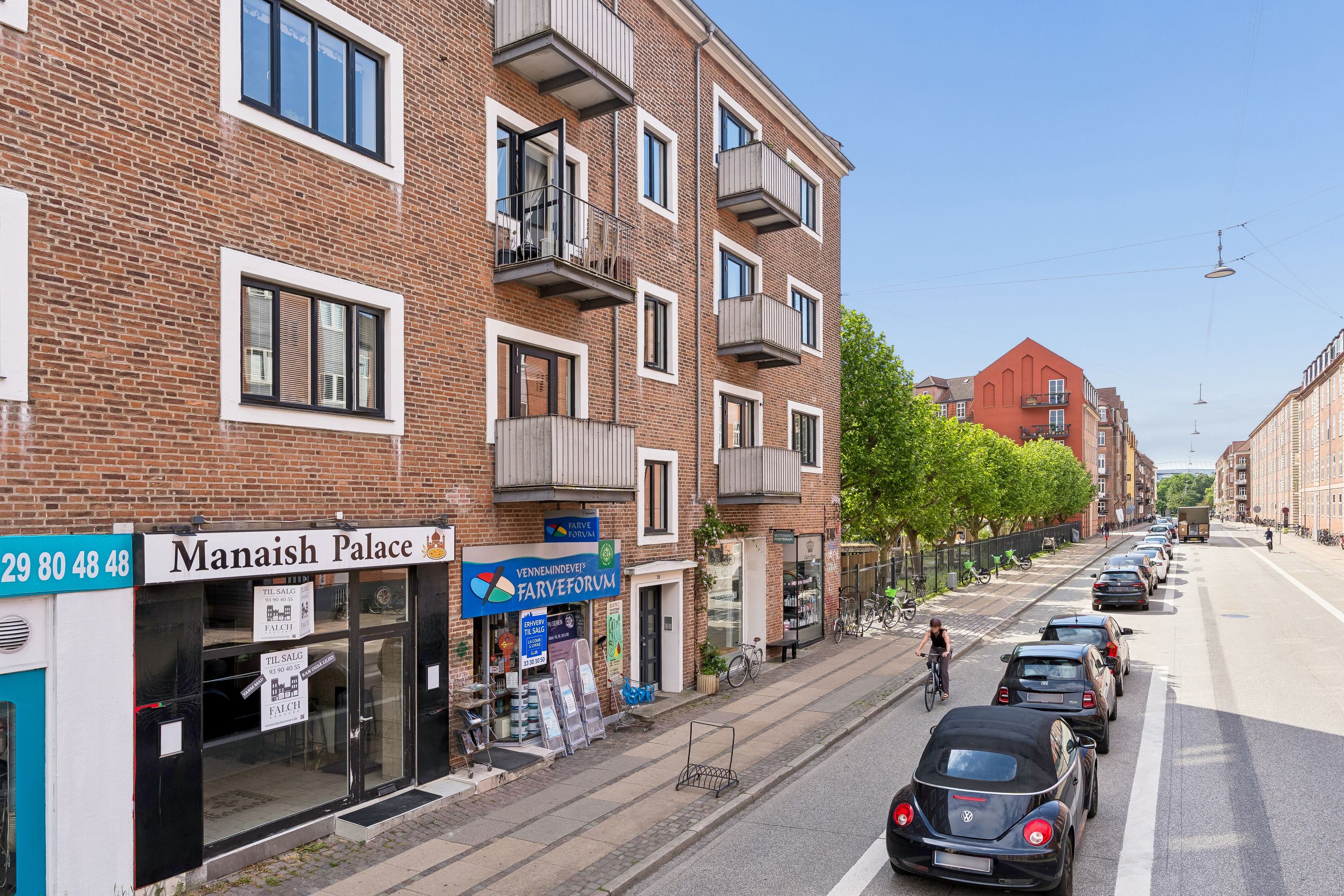 Biler parkeret på hver side af gaden med et kvindeligt cykelkøre bagved. Butikslokaler med skilte og mange cykler parkeret foran. En person kører en cykel langs siden af gaden