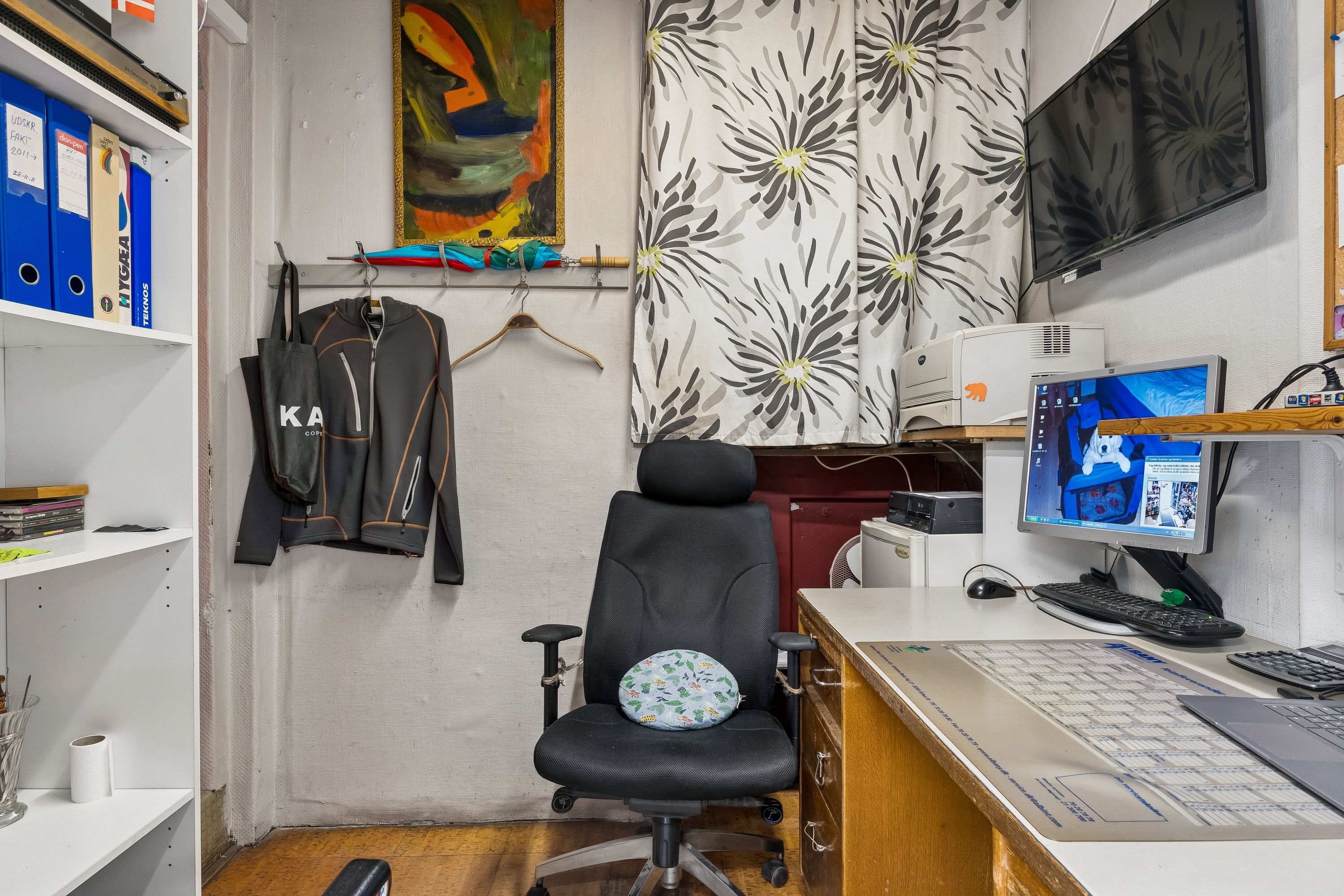 Kontor med computer, stol og hylde med mapper, papirkurv og briller. Væg med maleri, tøj og paraply. Skrivebord med printer og flere computere samt en tv-skærm.