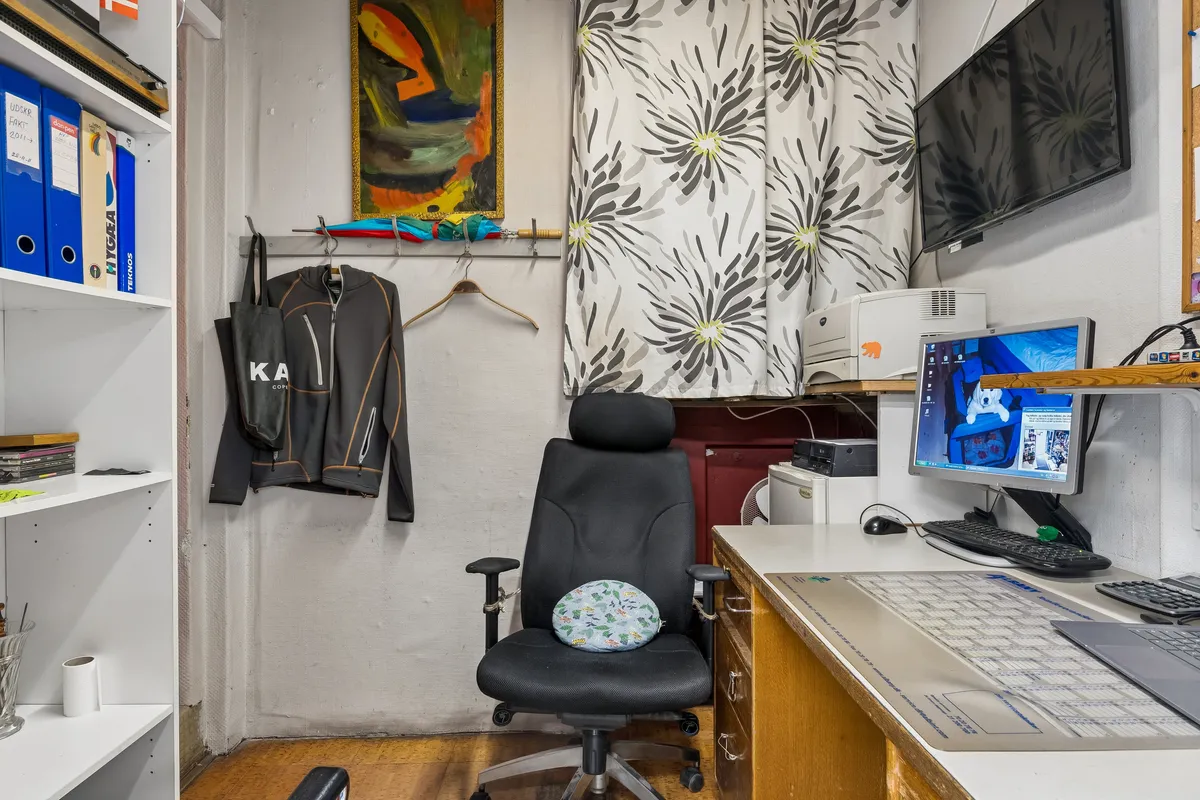Kontor med computer, stol og hylde med mapper, papirkurv og briller. Væg med maleri, tøj og paraply. Skrivebord med printer og flere computere samt en tv-skærm.