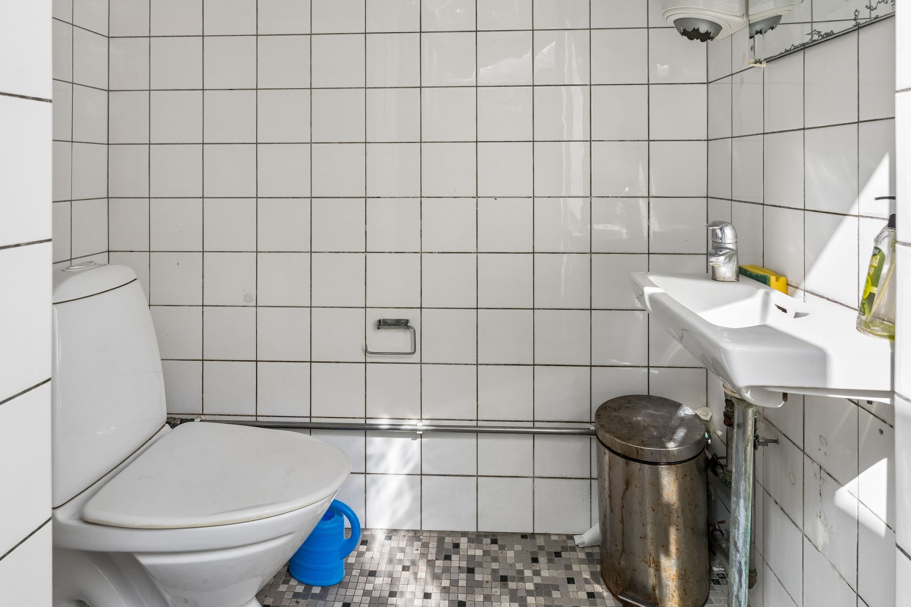 En hvid toilet i et lille badeværelse med toiletpapirholder og børste. Hvidt badeværelse med keramiske fliser og hvidt toilet. Et rørt badekar og et rustet metalaffaldsspand.