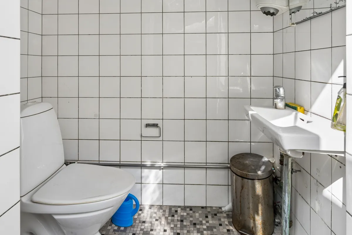 En hvid toilet i et lille badeværelse med toiletpapirholder og børste. Hvidt badeværelse med keramiske fliser og hvidt toilet. Et rørt badekar og et rustet metalaffaldsspand.
