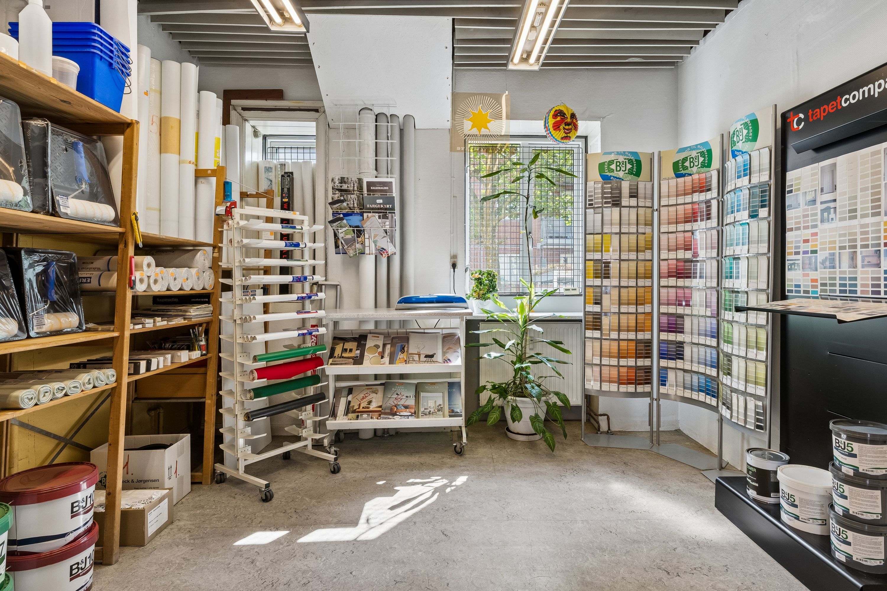 Maleributik med bjælkeloft og højt gulv. Forskellige farveprøver, tæpper og malingskanister i bunden. Planter og smykke på væggen
