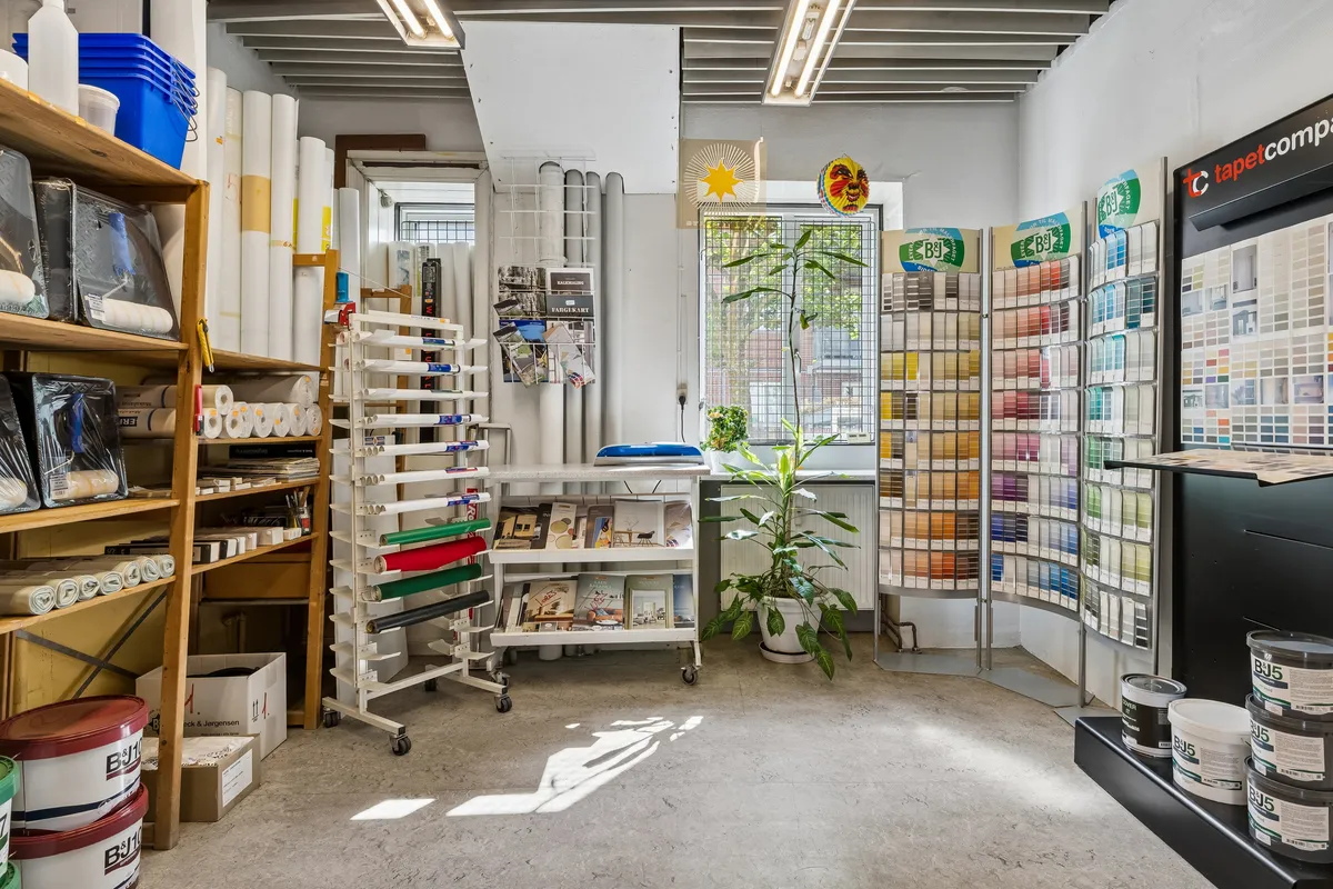 Maleributik med bjælkeloft og højt gulv. Forskellige farveprøver, tæpper og malingskanister i bunden. Planter og smykke på væggen