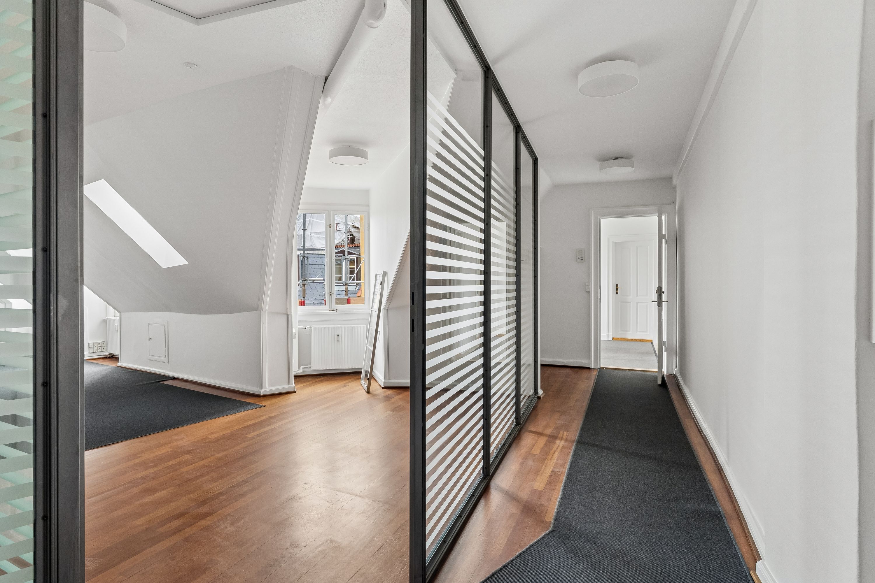 Kontor til leje med højloft, gulvtæppe og hvidt loft og vægge. Vindue med hvidt gardin og lyse skygger på loftet. Glasskive med stribet mønster og lysepunkter.