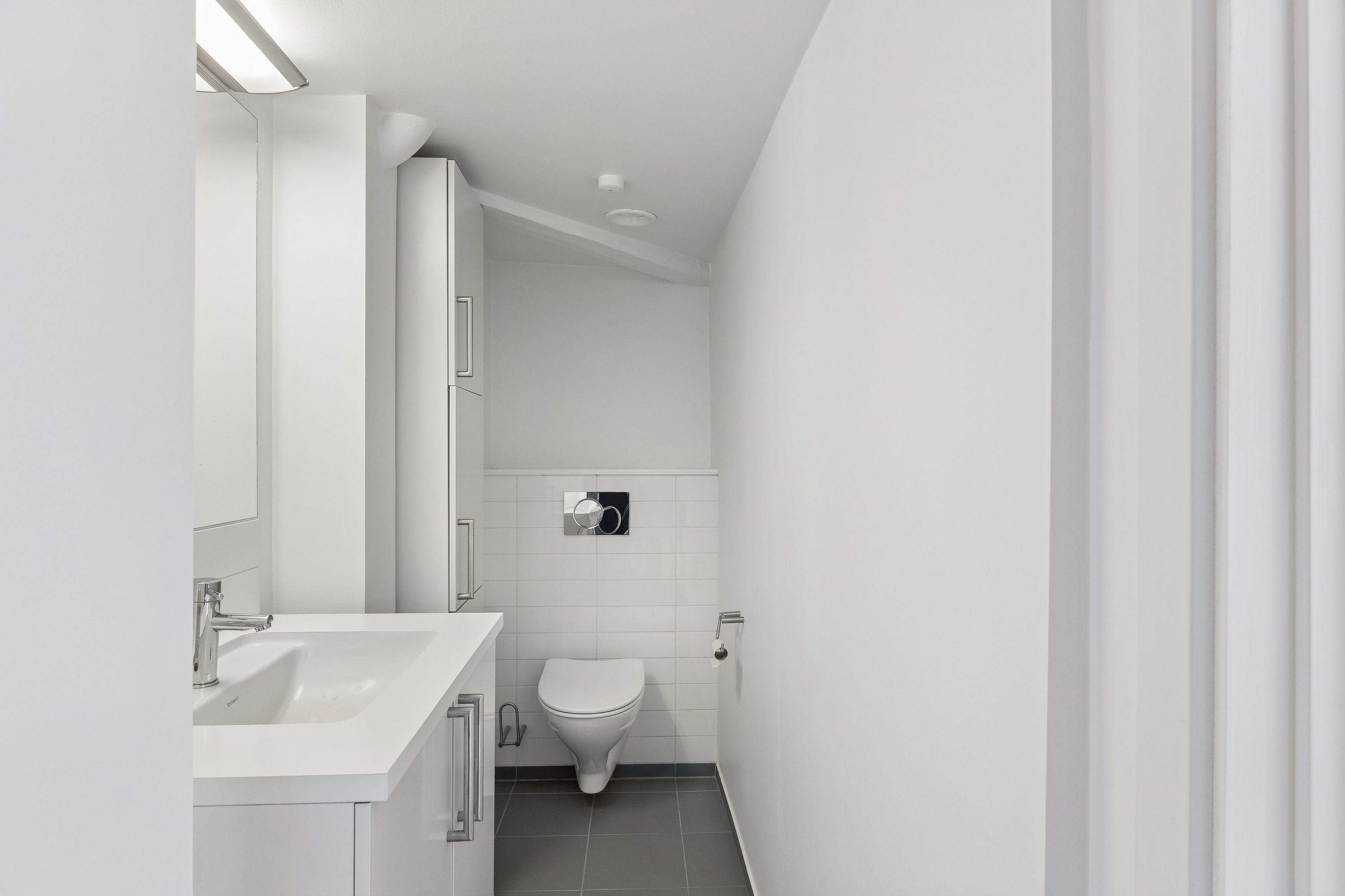 En moderne toiletrum med hvidt loft, vægge og gulv, hvidt toilet, hvidt vask og en høj hvid toiletkabinet samt hvide håndtagsgreb